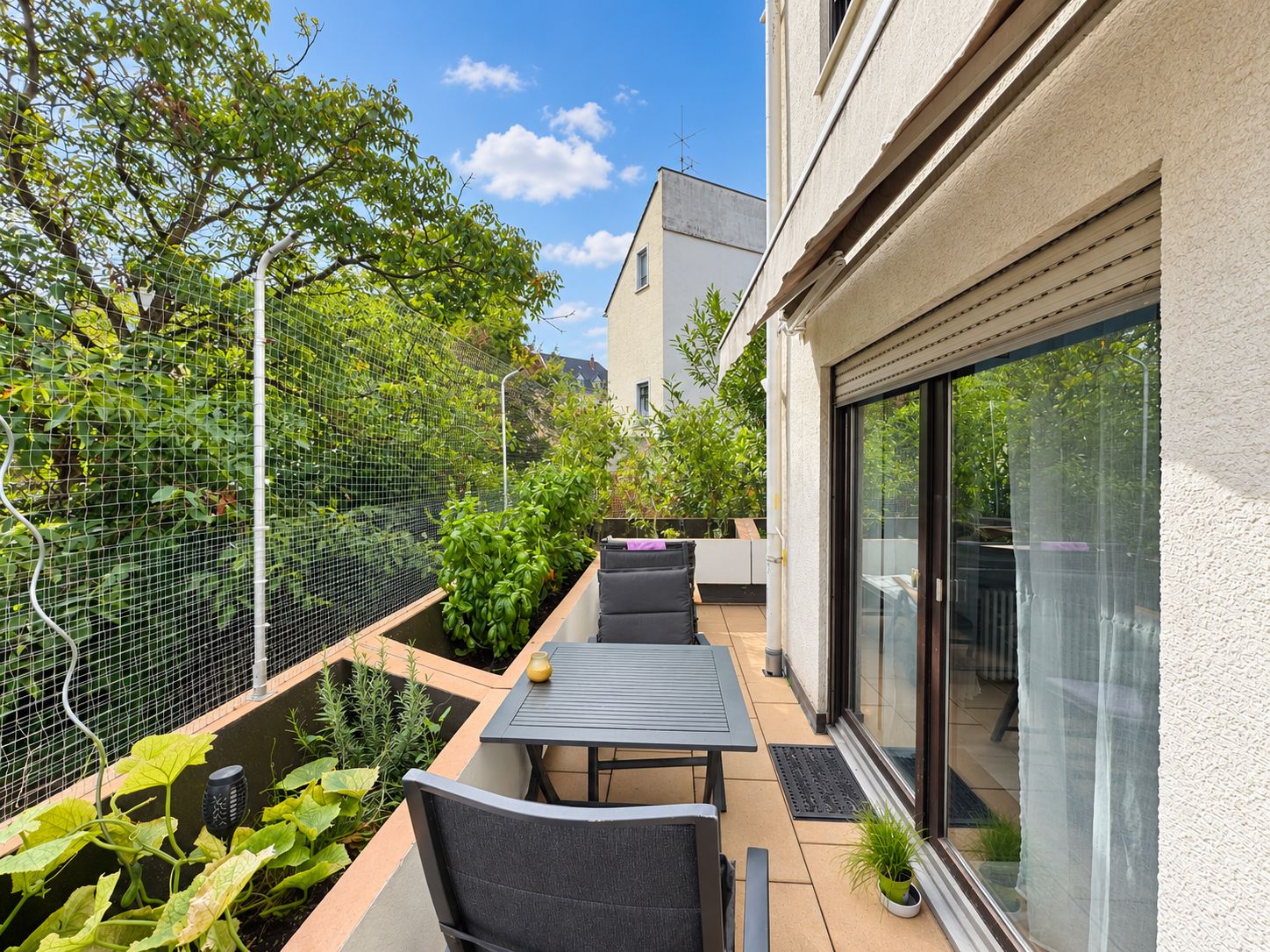 Balkon mit Tisch, zwei Stühlen und Pflanzen neben einem Wohnhaus an einem sonnigen Tag.