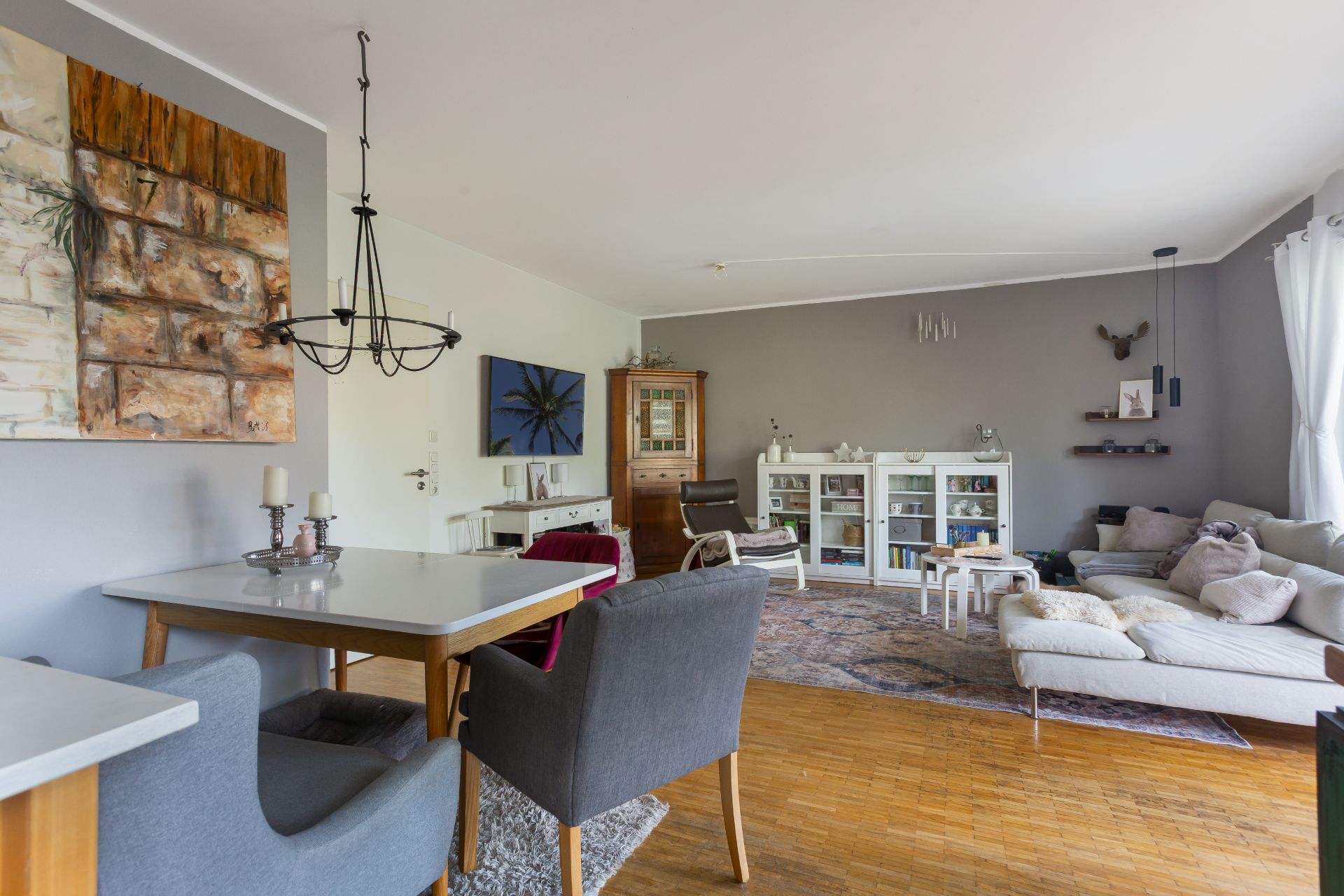 Helles Wohnzimmer mit Esstisch, grauen Sesseln, Sofa, Bücherregalen und großen Fenstern mit weißen Vorhängen.