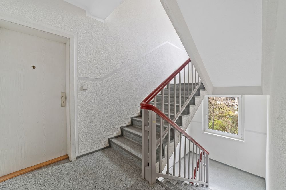 Helles Treppenhaus mit geschlossener Wohnungstür und Fenster mit Blick nach draußen.