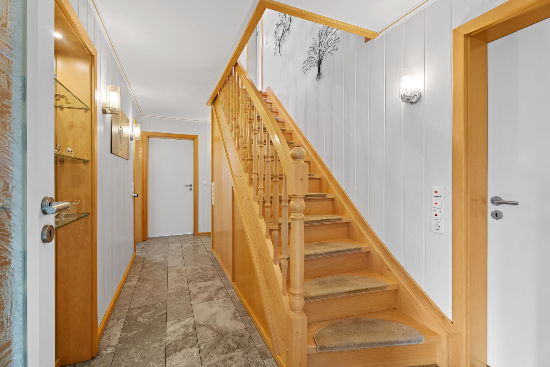 Heller Flur mit Holztreppe, mehreren Türen und Wanddekoration aus Metall.