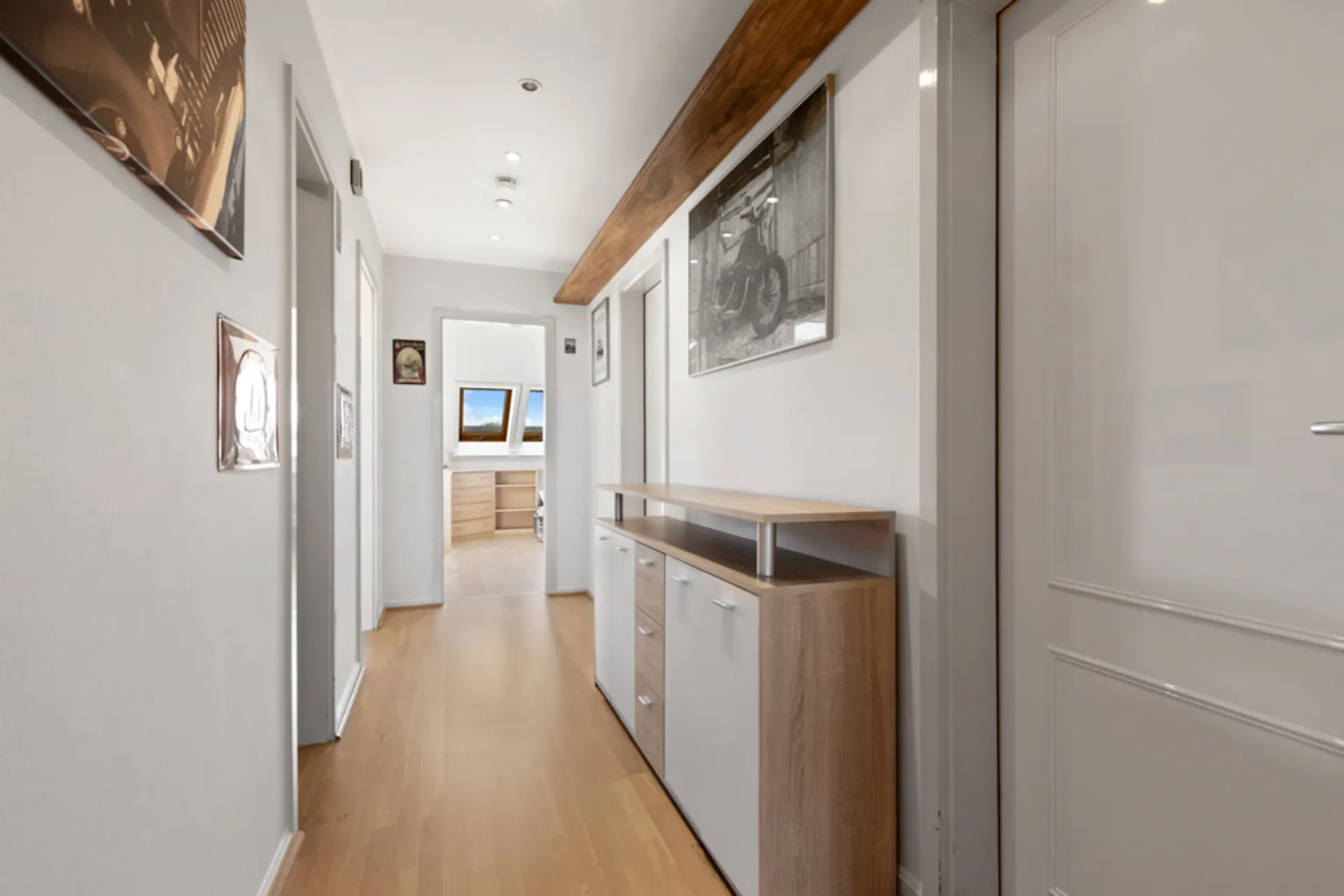 Heller Flur mit Holzfußboden, Kommode und Wandbildern, im Hintergrund ein Zimmer mit Dachfenstern.