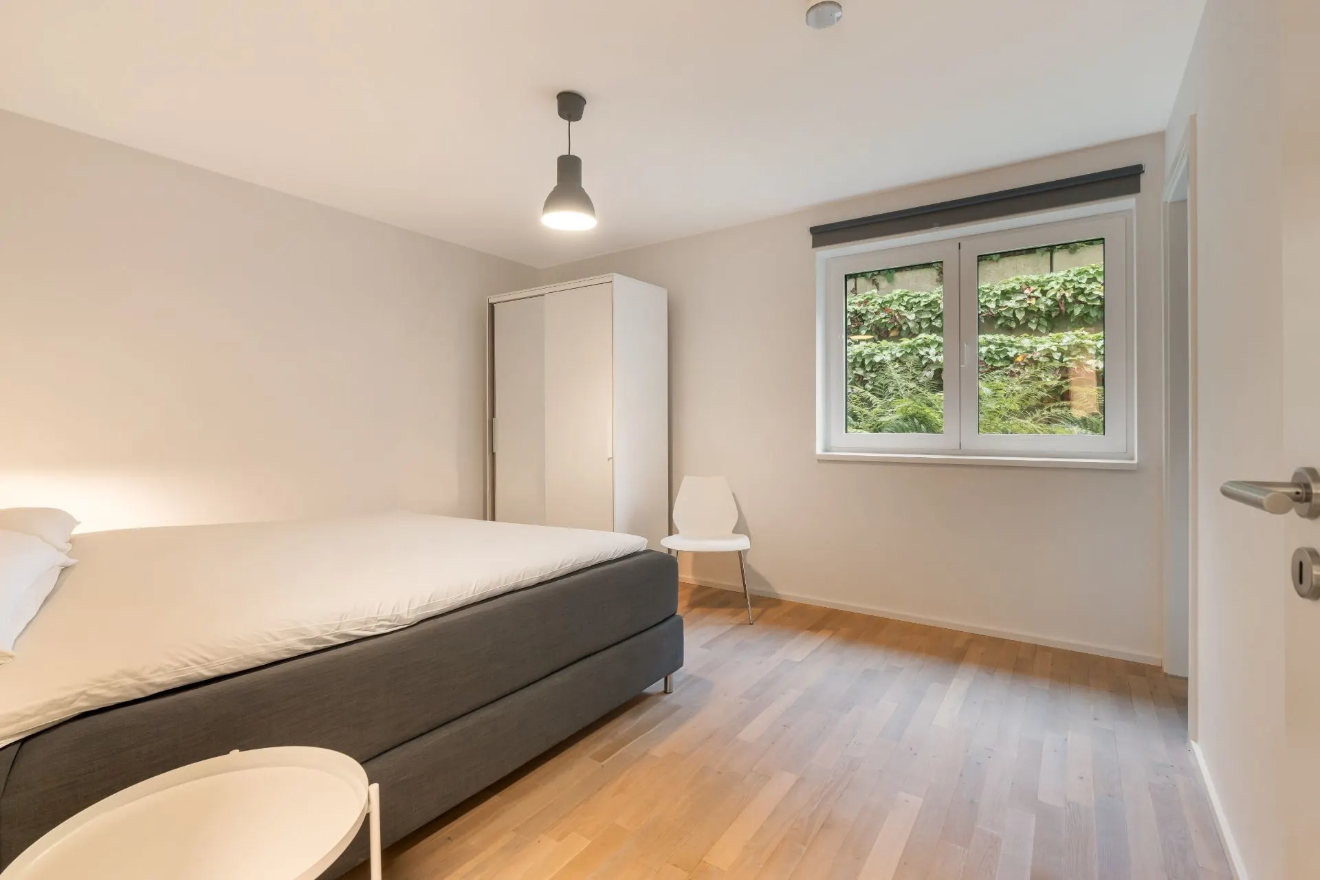 Helles Schlafzimmer mit Bett, Kleiderschrank, Stuhl, Beistelltisch und Fenster mit Blick ins Grüne.