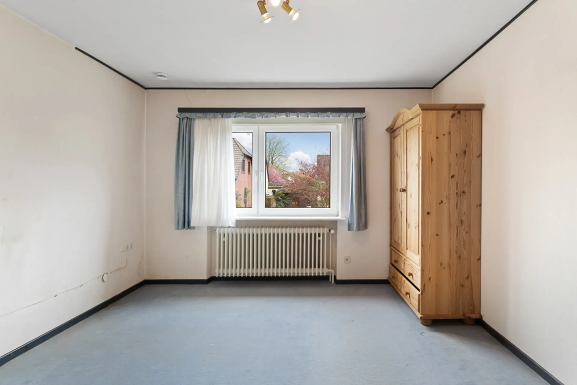 Helles Zimmer mit Fenster, Heizung, Vorhängen und einem Kleiderschrank aus Holz.
