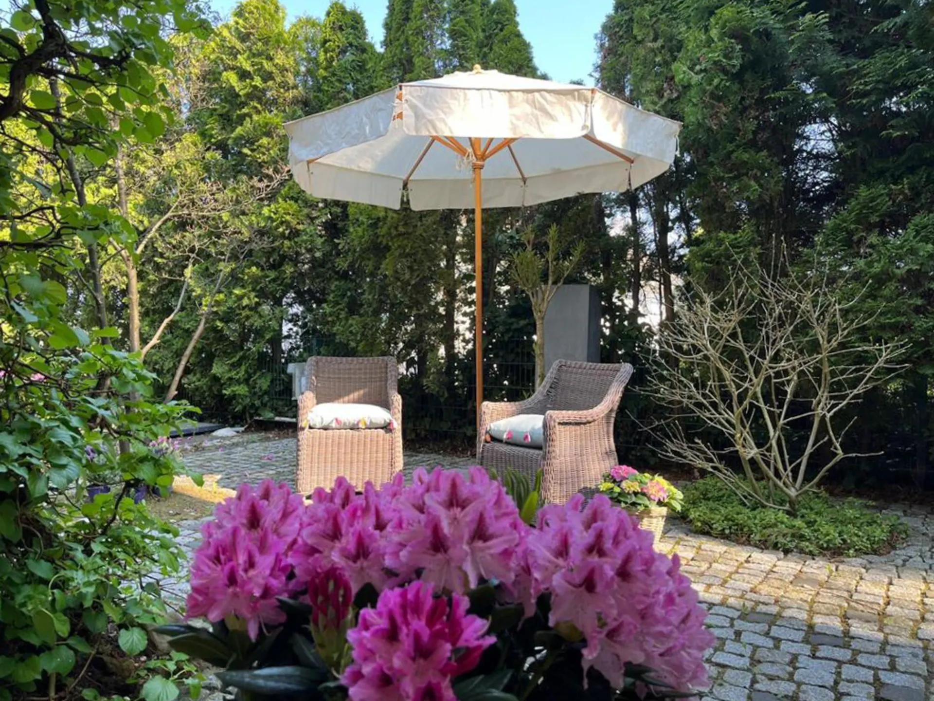 Zwei Korbstühle und ein Sonnenschirm auf einer Terrasse mit Blumen und viel Grün.