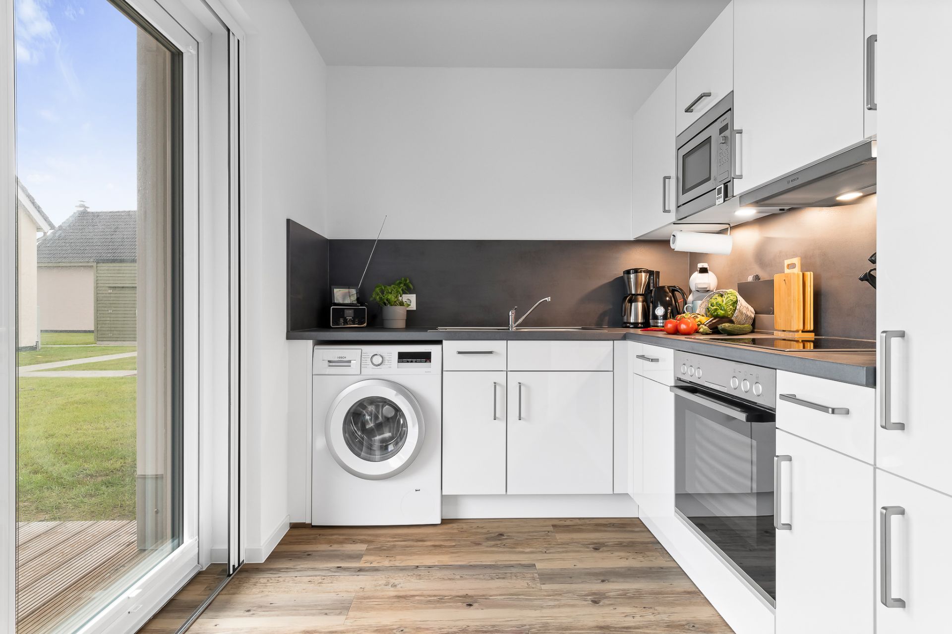 Moderne weiße Einbauküche mit Waschmaschine, Backofen und großen Fenstern mit Blick nach draußen.