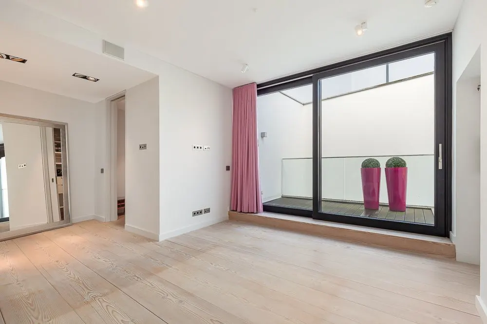 Helles Zimmer mit Holzfußboden, Glasfront, rosa Vorhang und Balkon mit zwei pinken Pflanzgefäßen.