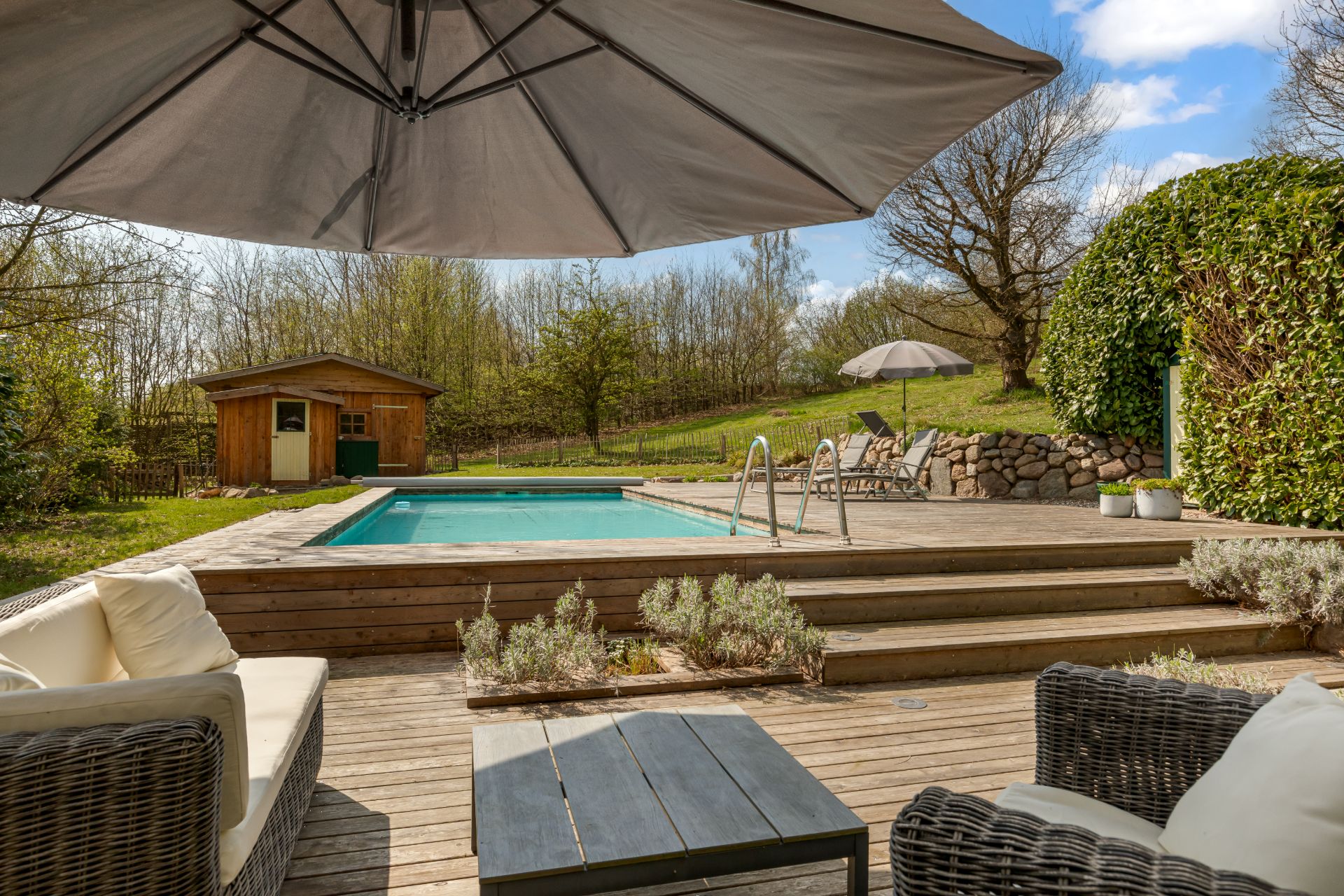 Terrasse mit Sitzgruppe, großem Sonnenschirm, Pool, Stufen und Gartenhaus im Grünen.