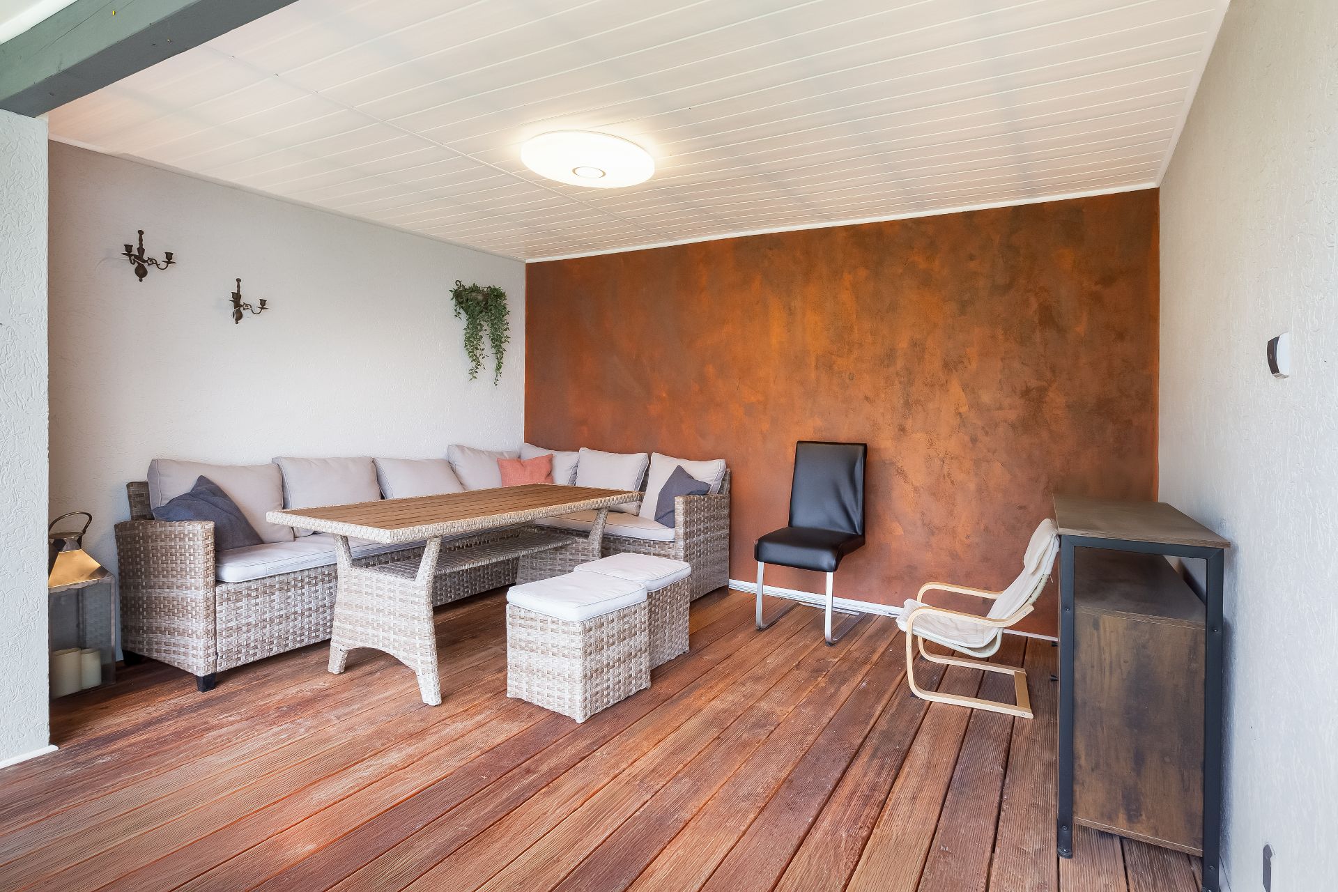 Überdachte Terrasse mit Sitzgruppe, Holztisch, zwei Stühlen und Sideboard auf Holzboden.