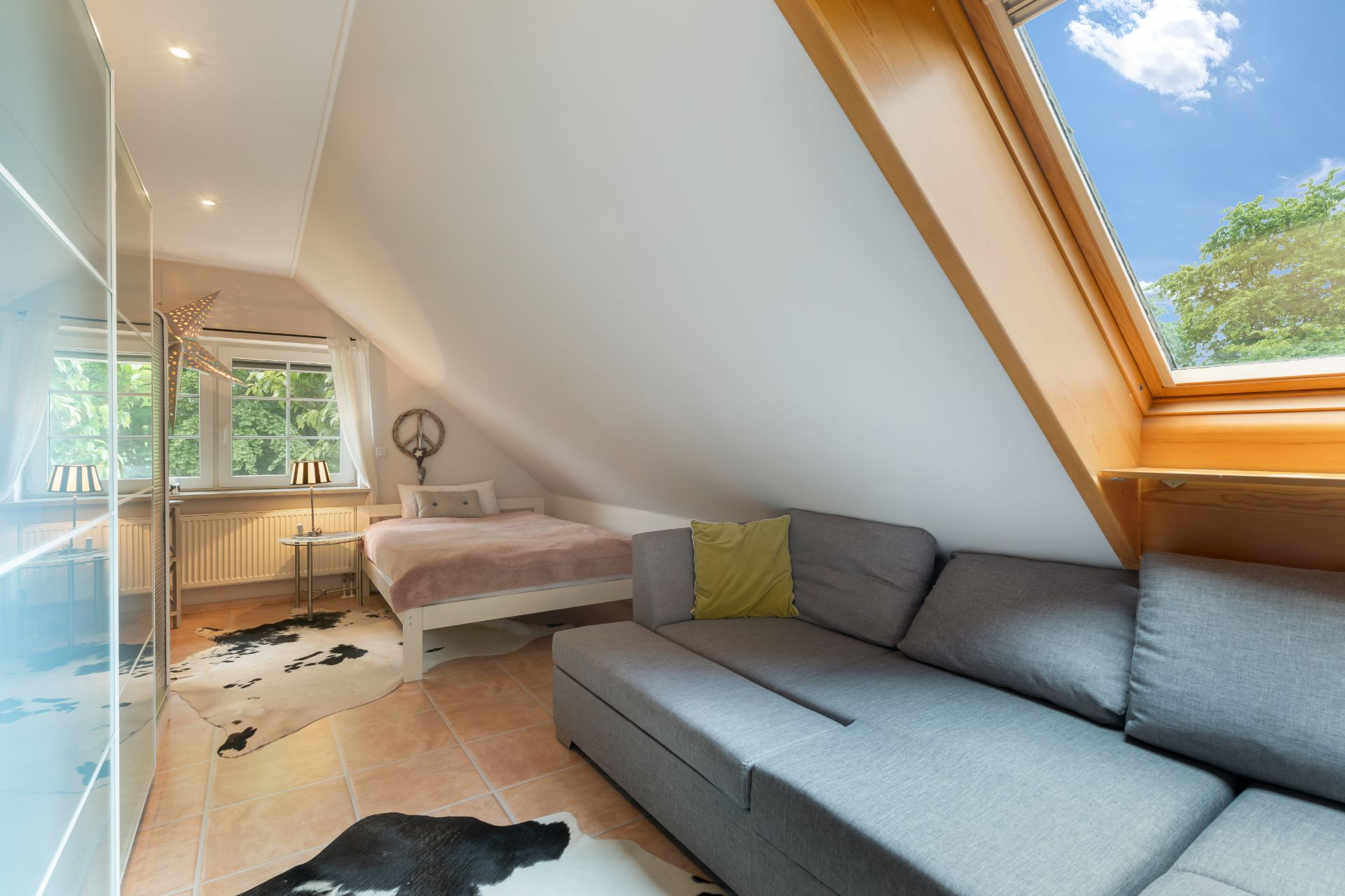 Wohnzimmer mit grauem Sofa, Bett, Fenster und Dachfenster unter einer Schräge.