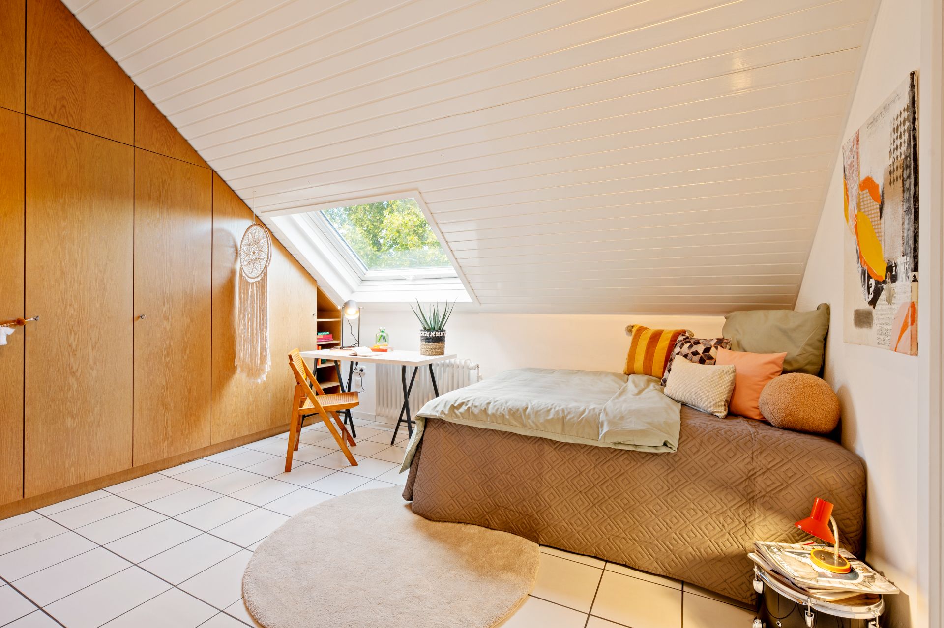 Helles Schlafzimmer mit Dachschräge, Bett, Schreibtisch am Fenster und Holzeinbauschränken.