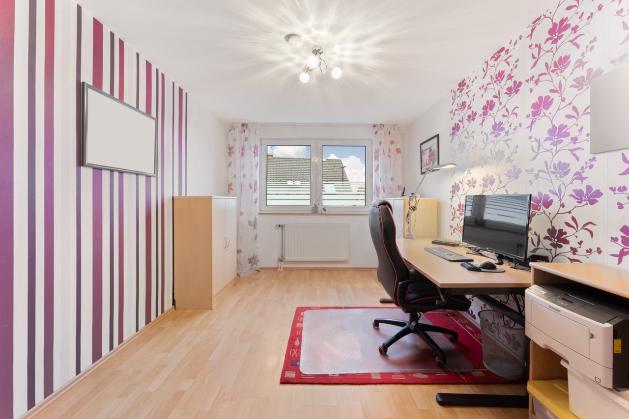 Arbeitszimmer mit Schreibtisch, Stuhl, Computer und bunter Tapete, großes Fenster im Hintergrund.