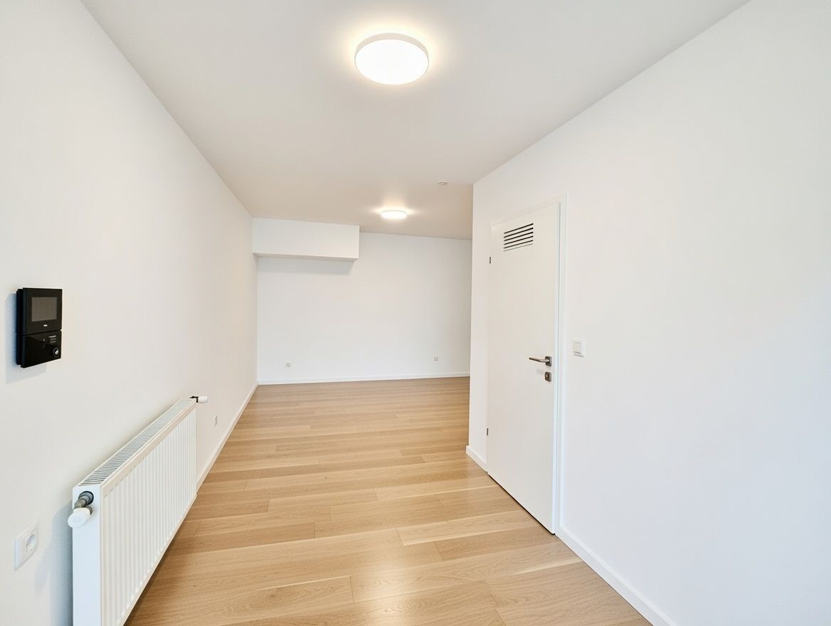 Leerer, heller Raum mit Holzboden, weißen Wänden, Heizkörper und geschlossener Tür.