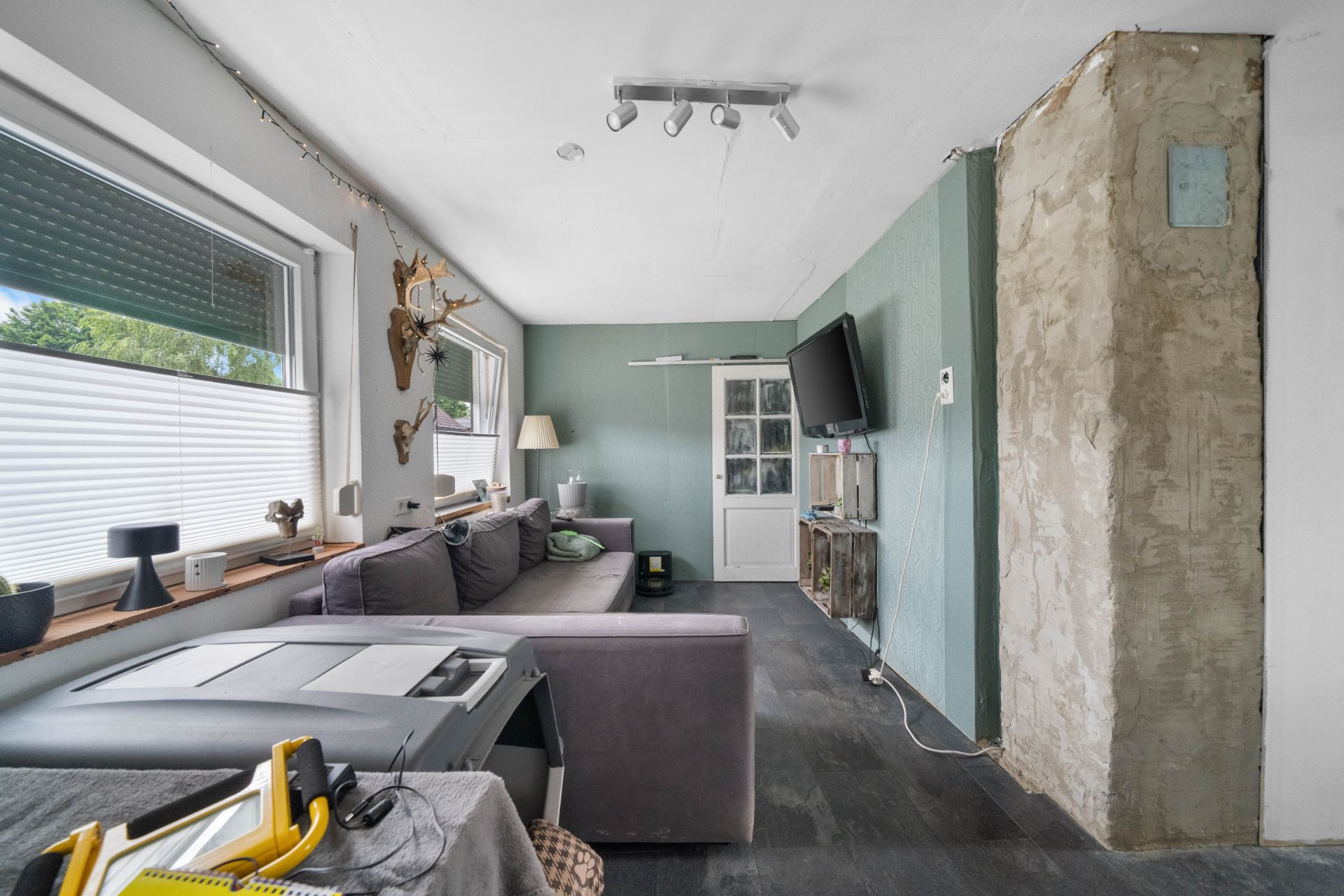 Wohnzimmer mit grauem Sofa, Wand-TV, Fenstern und teilweise verputzter Wand.