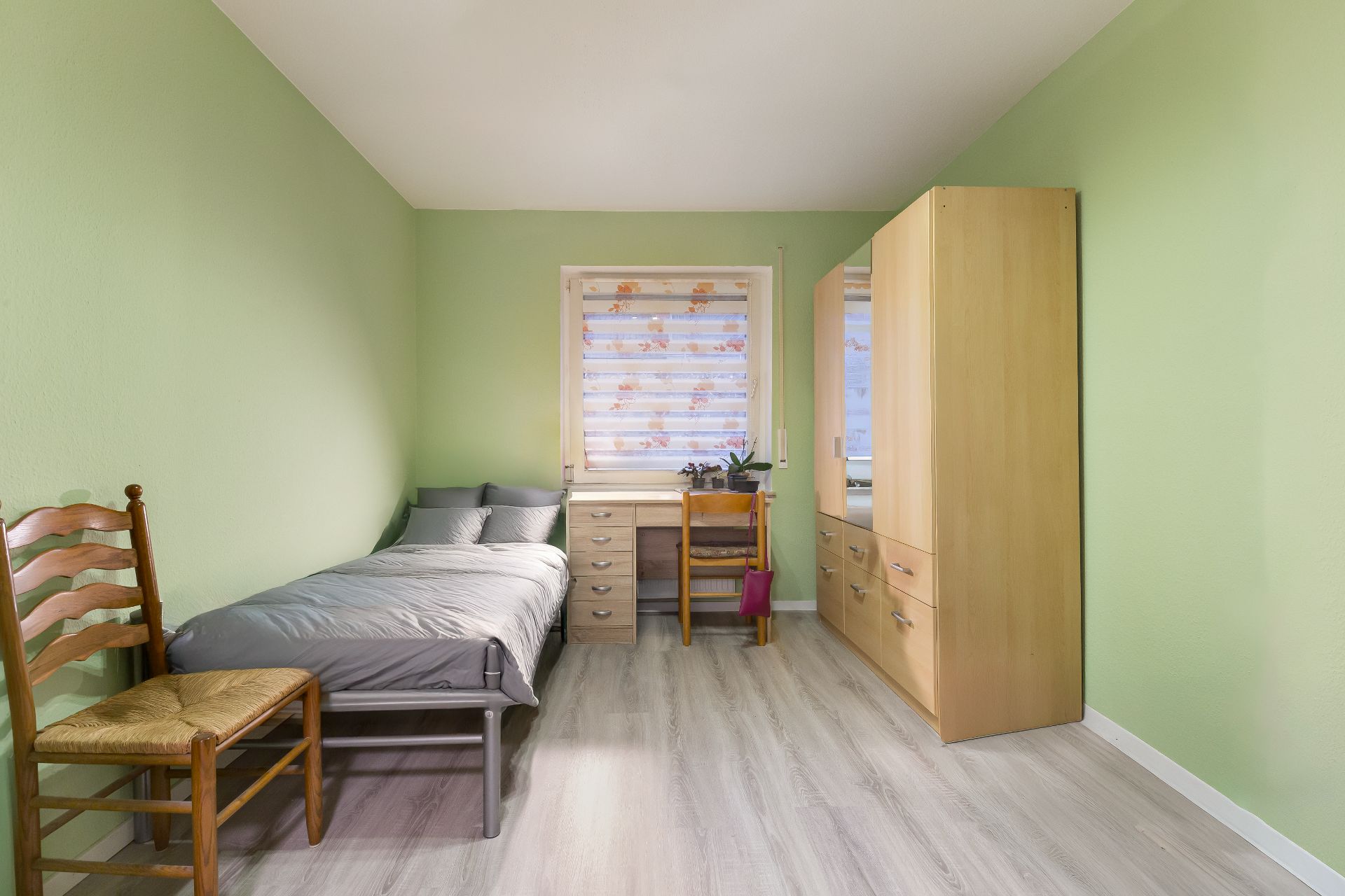 Ein schlichtes Schlafzimmer mit Einzelbett, Holzstuhl, Schreibtisch und Kleiderschrank.