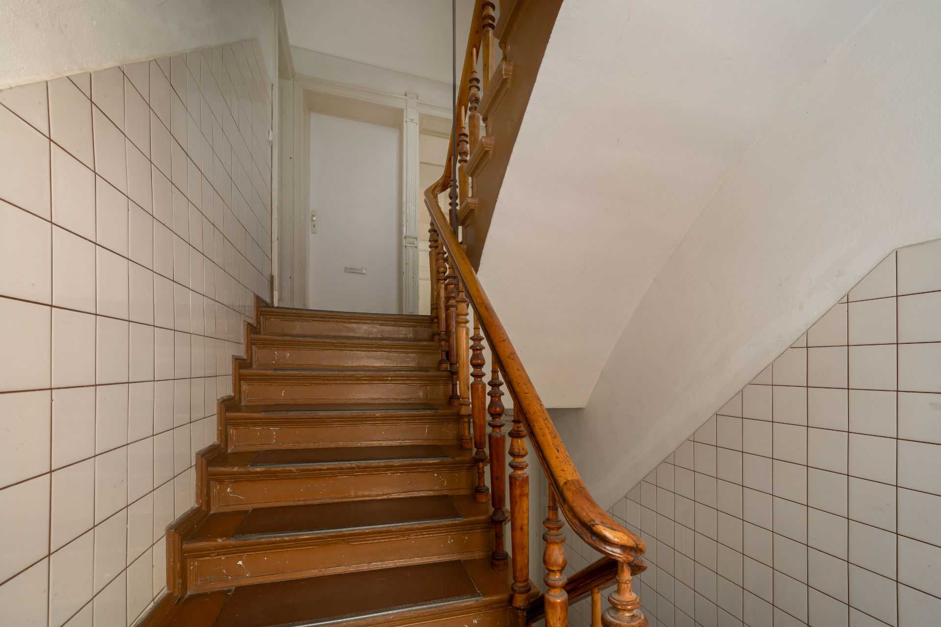 Braune Holztreppe mit Geländer und weißen gekachelten Wänden in einem Wohnhaus.