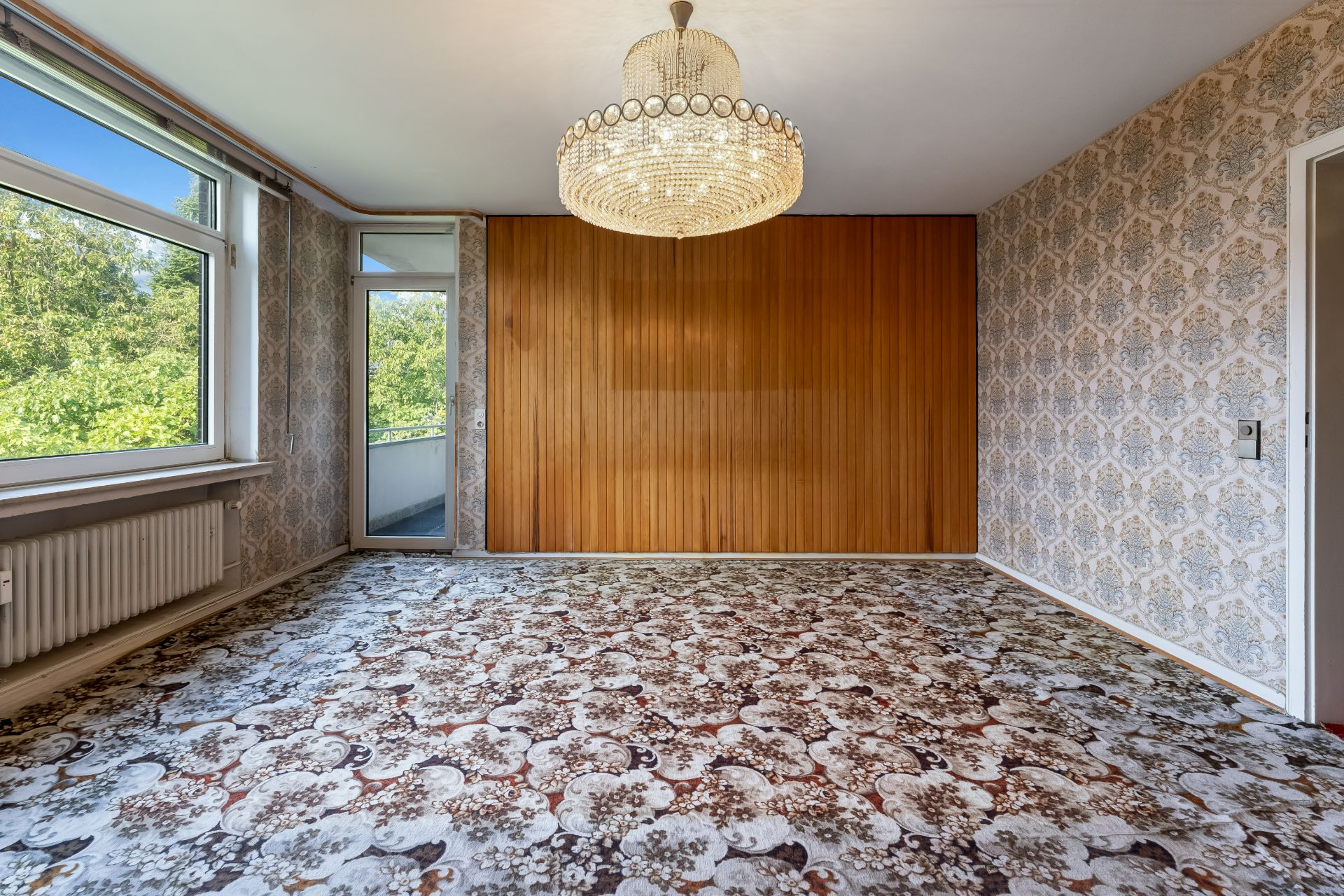 Leeres Zimmer mit gemustertem Teppich, großem Fenster, Holzwand und Kronleuchter.