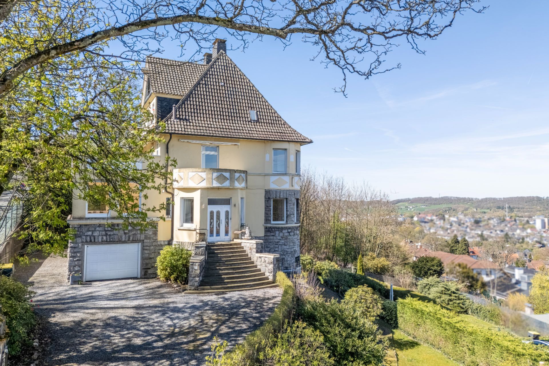Großes Wohnhaus mit Steintreppe, Garage und Garten, im Hintergrund Blick auf Stadtlandschaft.