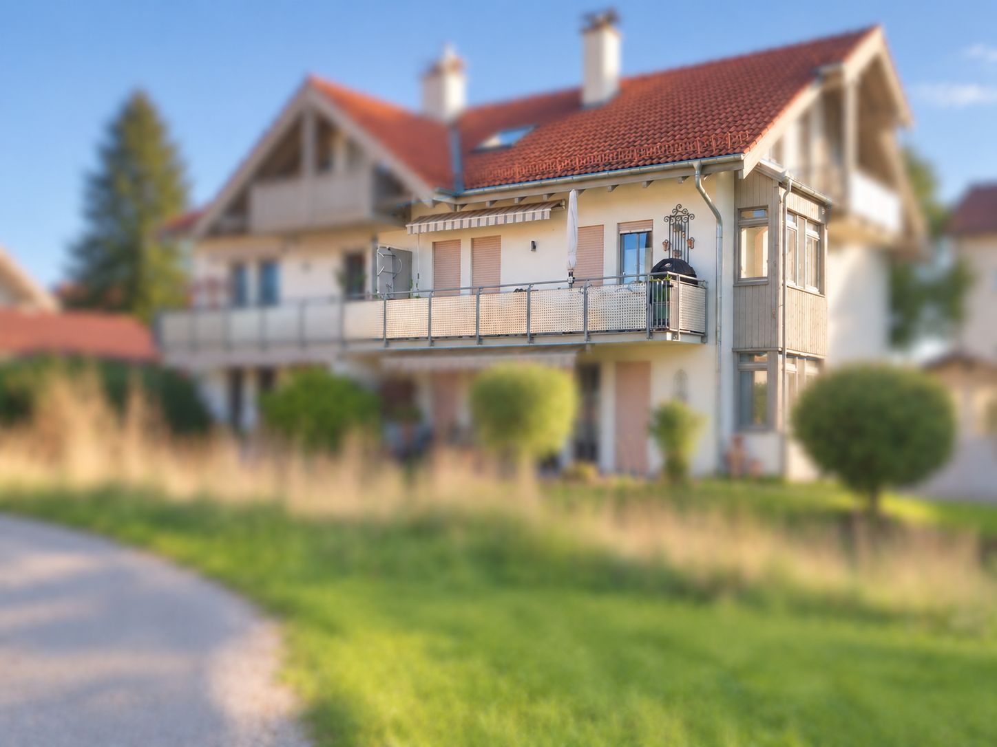Wohnhaus mit rotem Dach, großem Balkon und gepflegtem Garten bei sonnigem Wetter.