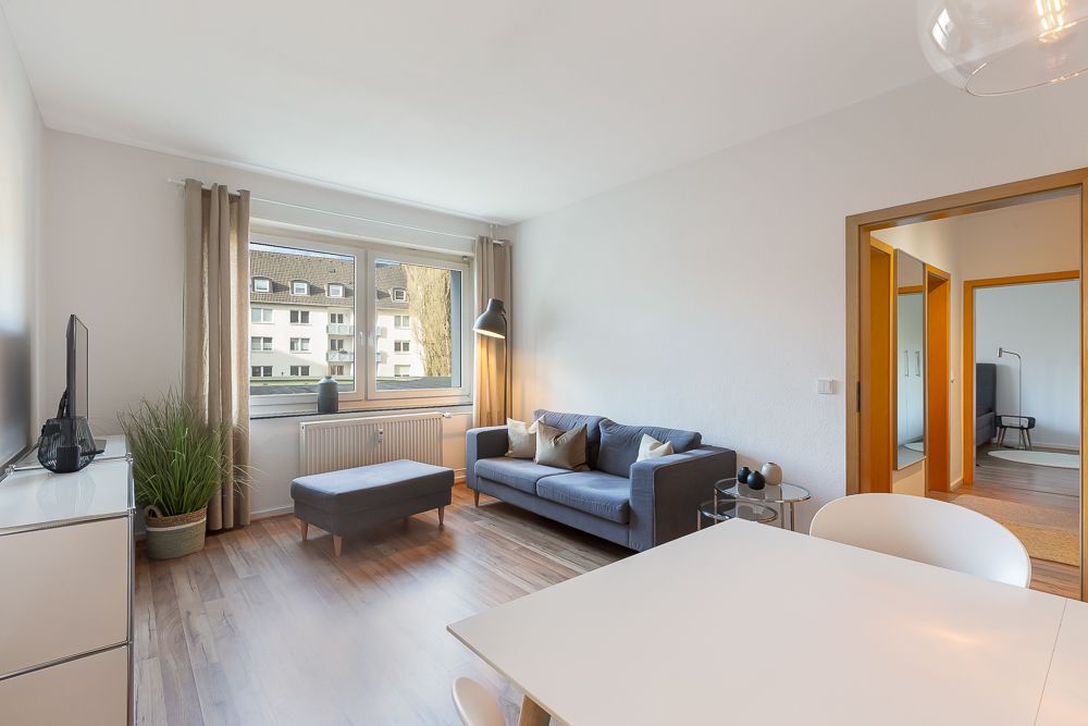 Helles Wohnzimmer mit grauem Sofa, kleinem Tisch, Ottomane und großem Fenster mit Vorhängen.