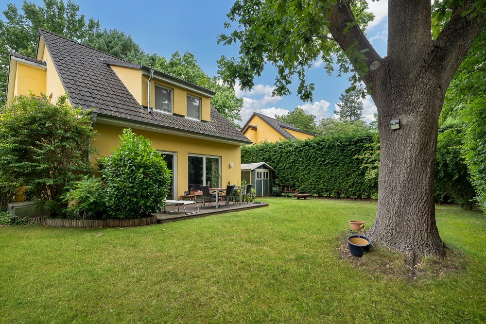 Garten mit großer Terrasse, gelbem Haus, Rasenfläche, Baum und Gartenhäuschen.