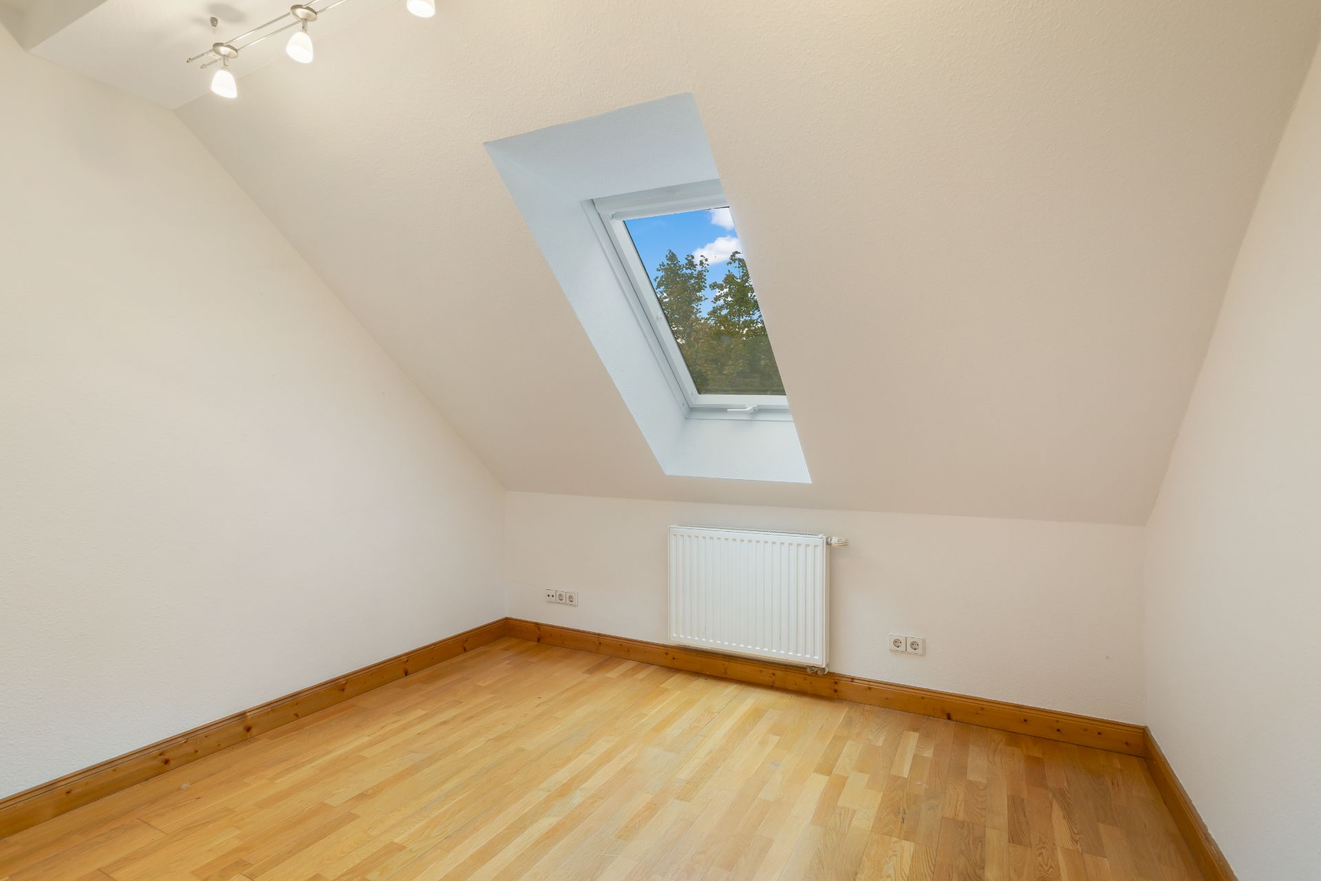 Leerer Raum mit Dachschräge, Holzboden, Dachfenster und Heizkörper an der Wand.