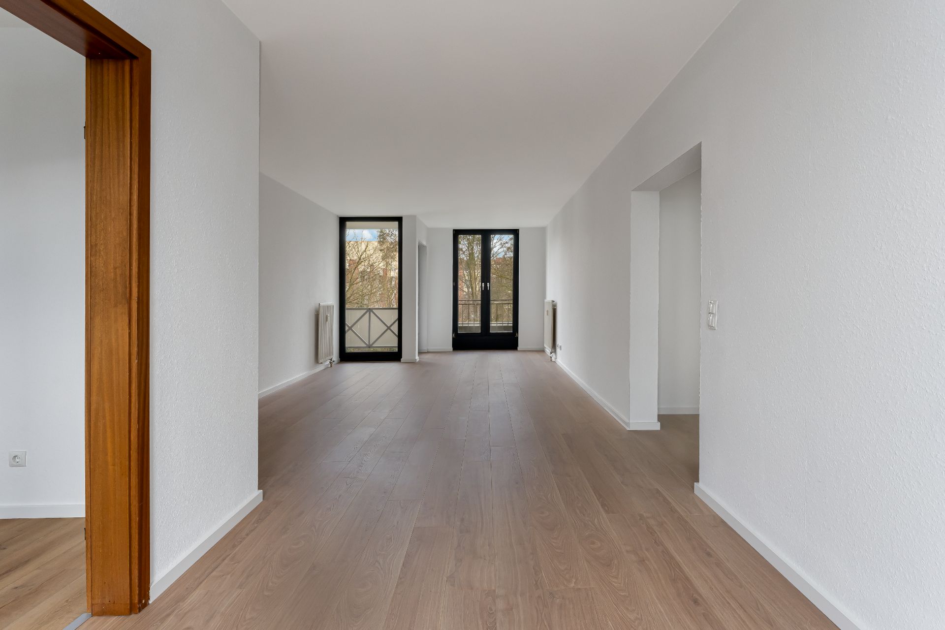 Leeres Wohnzimmer mit Holzboden, weißen Wänden und zwei großen Fenstern mit schwarzem Rahmen.