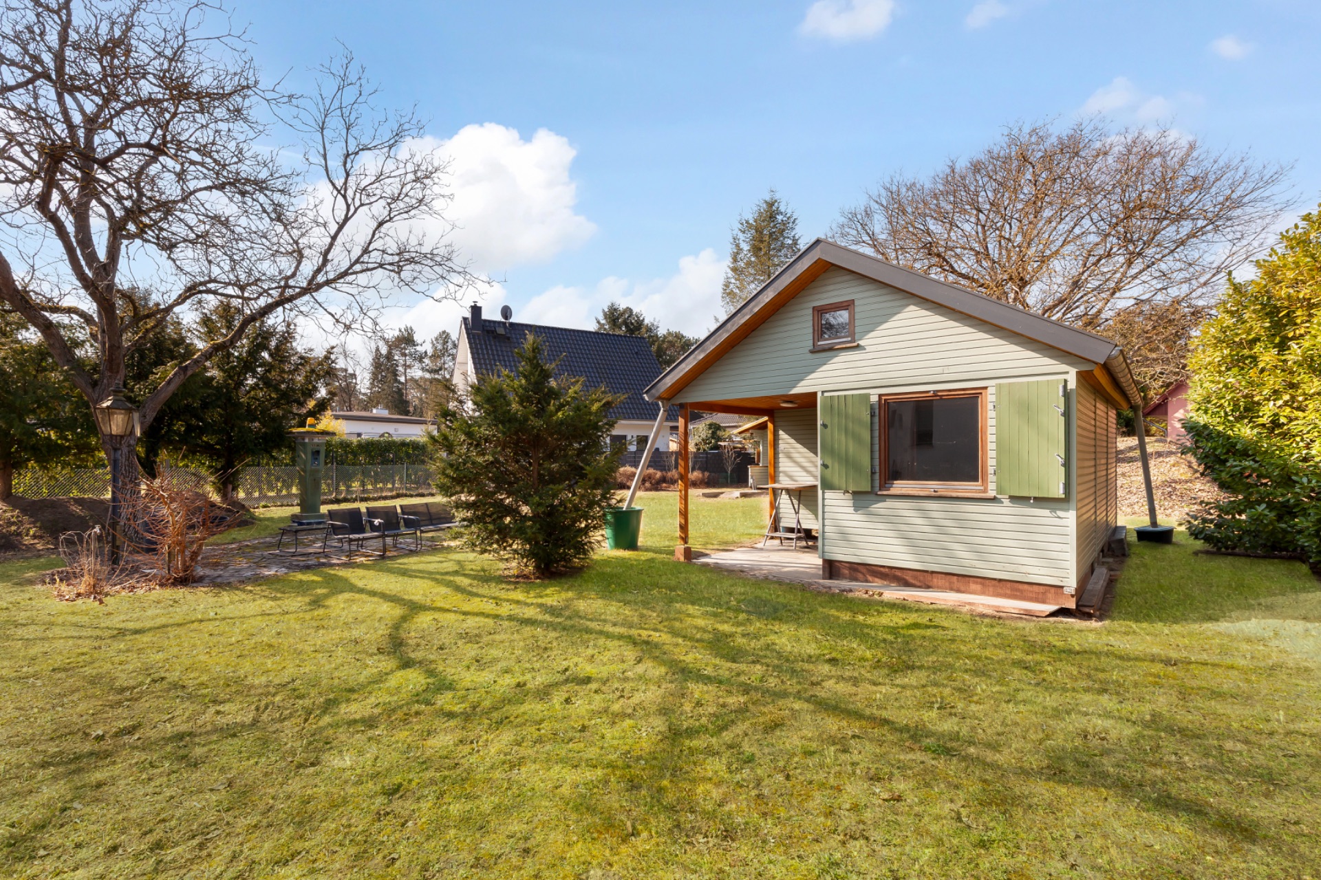 Kleines Gartenhaus mit Veranda auf einer grünen Wiese, umgeben von Bäumen und Sträuchern.