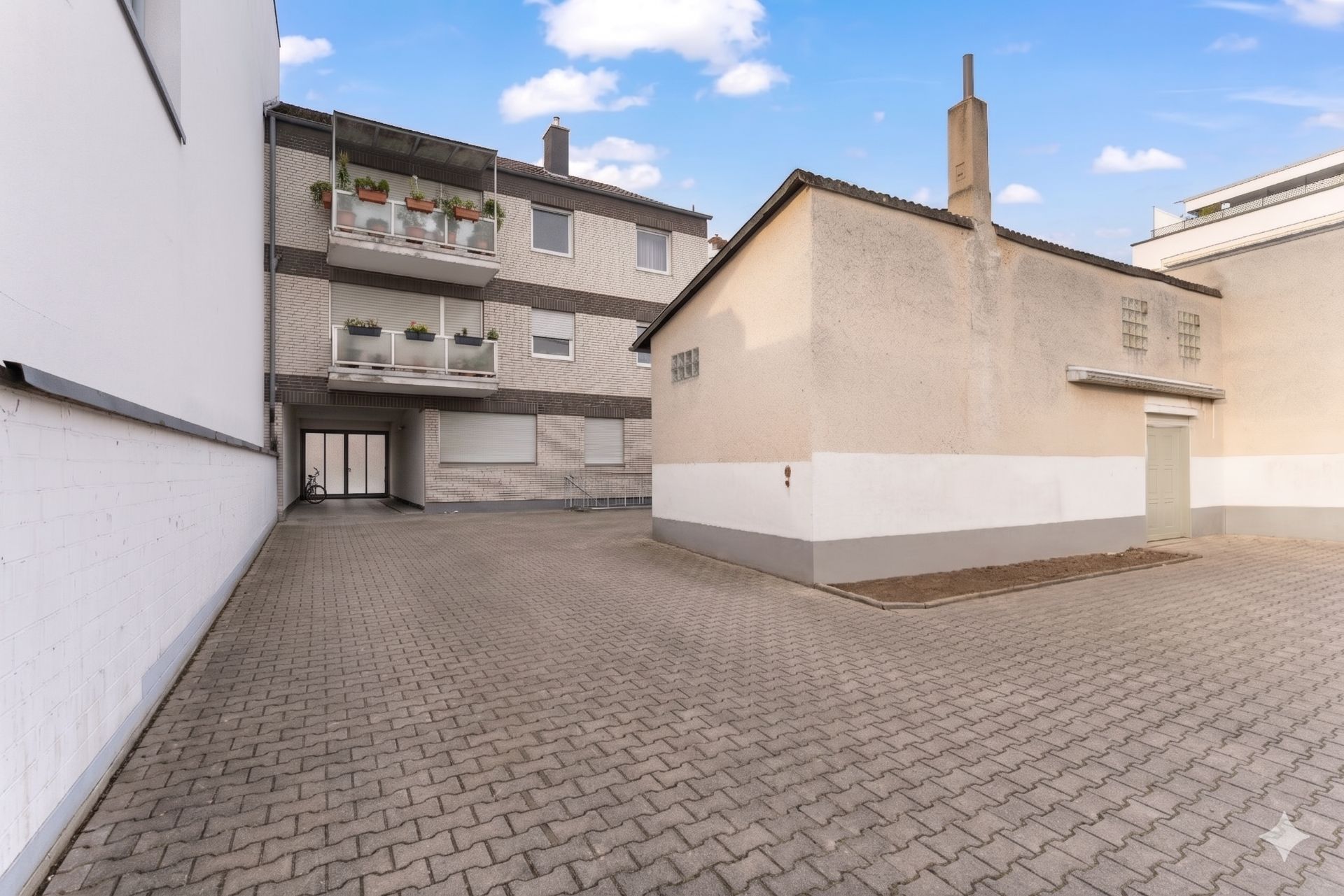 Gepflasterter Hof mit kleinem Nebengebäude und Wohnhaus mit Balkonen im Hintergrund.