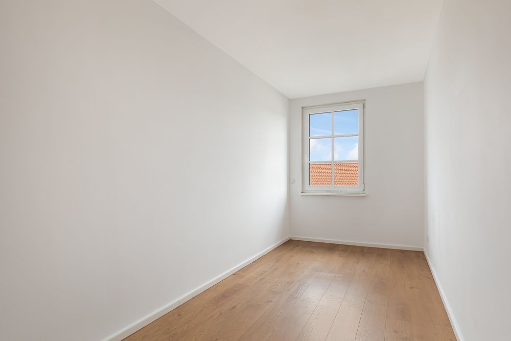 Leerer, schmaler Raum mit weiß gestrichenen Wänden, Holzfußboden und einem Fenster am Ende.