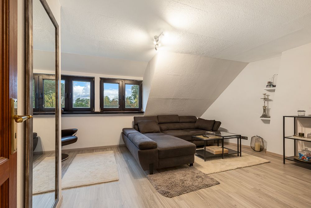 Wohnzimmer mit Ecksofa, Couchtisch, Regalen und drei Fenstern unter einer Dachschräge.