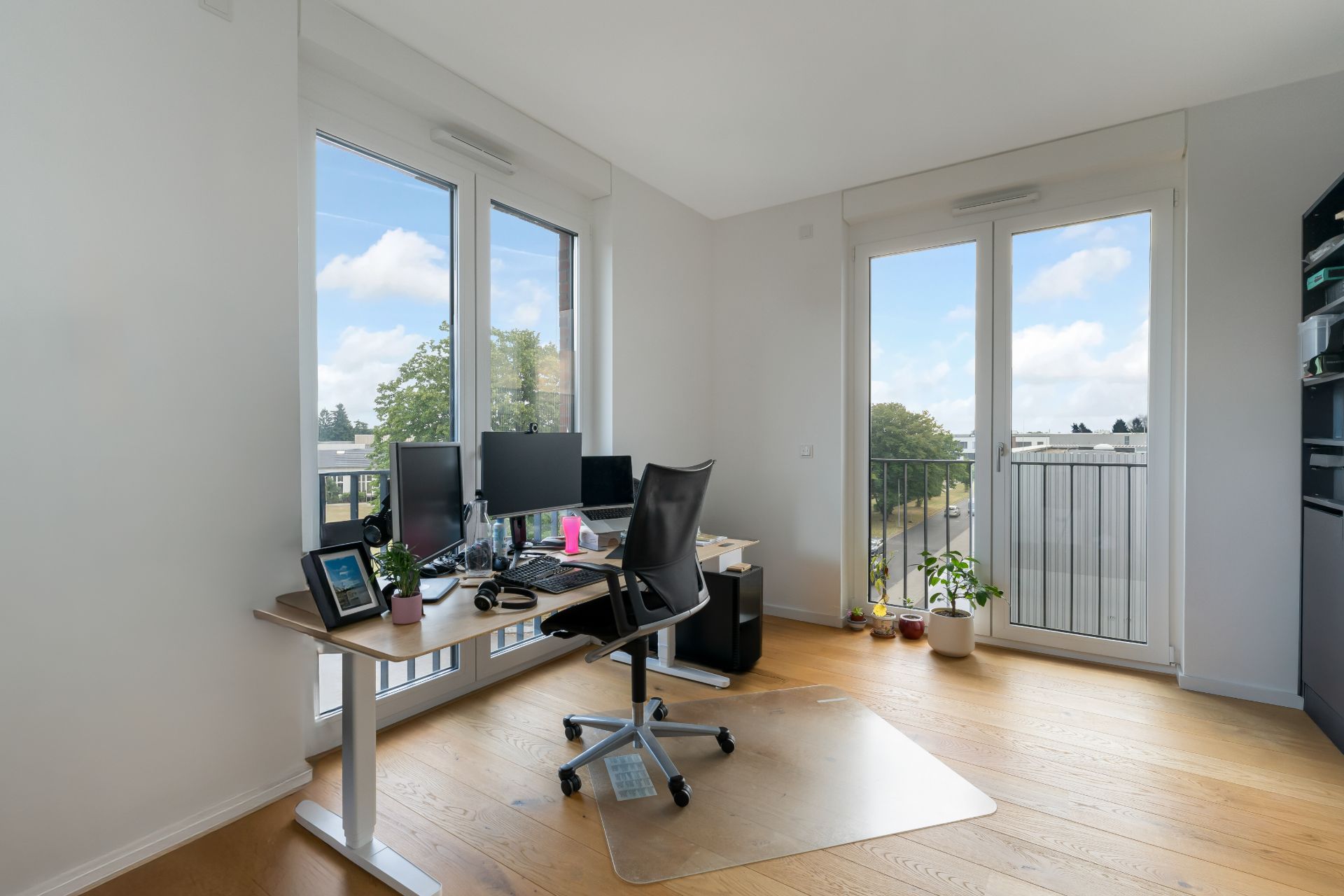 Helles Arbeitszimmer mit großem Schreibtisch, Monitoren und Pflanzen, Parkettboden, Fenster mit Ausblick.