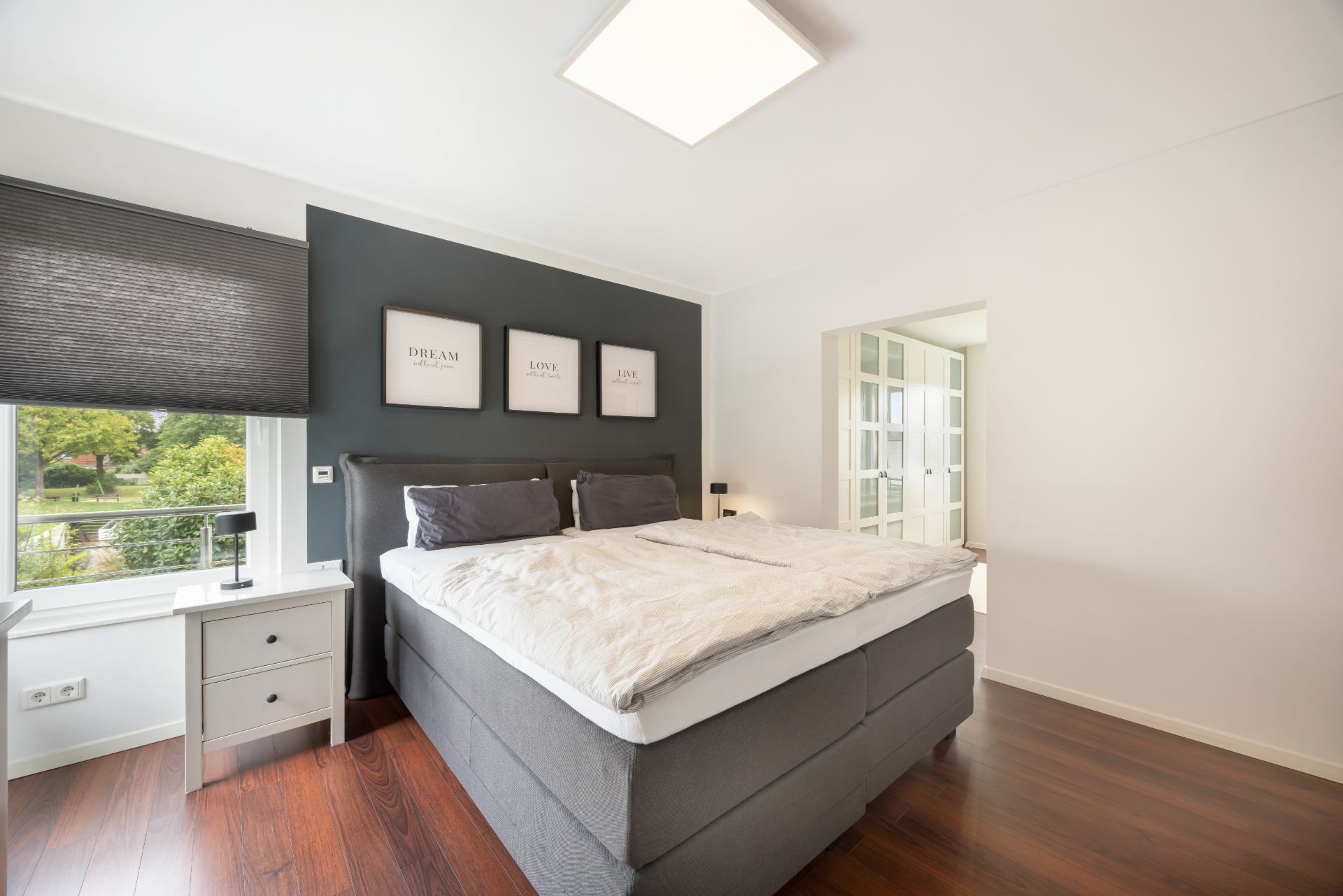 Modernes Schlafzimmer mit Doppelbett, dunklem Boden, Nachttisch und Fenster mit Blick ins Grüne.
