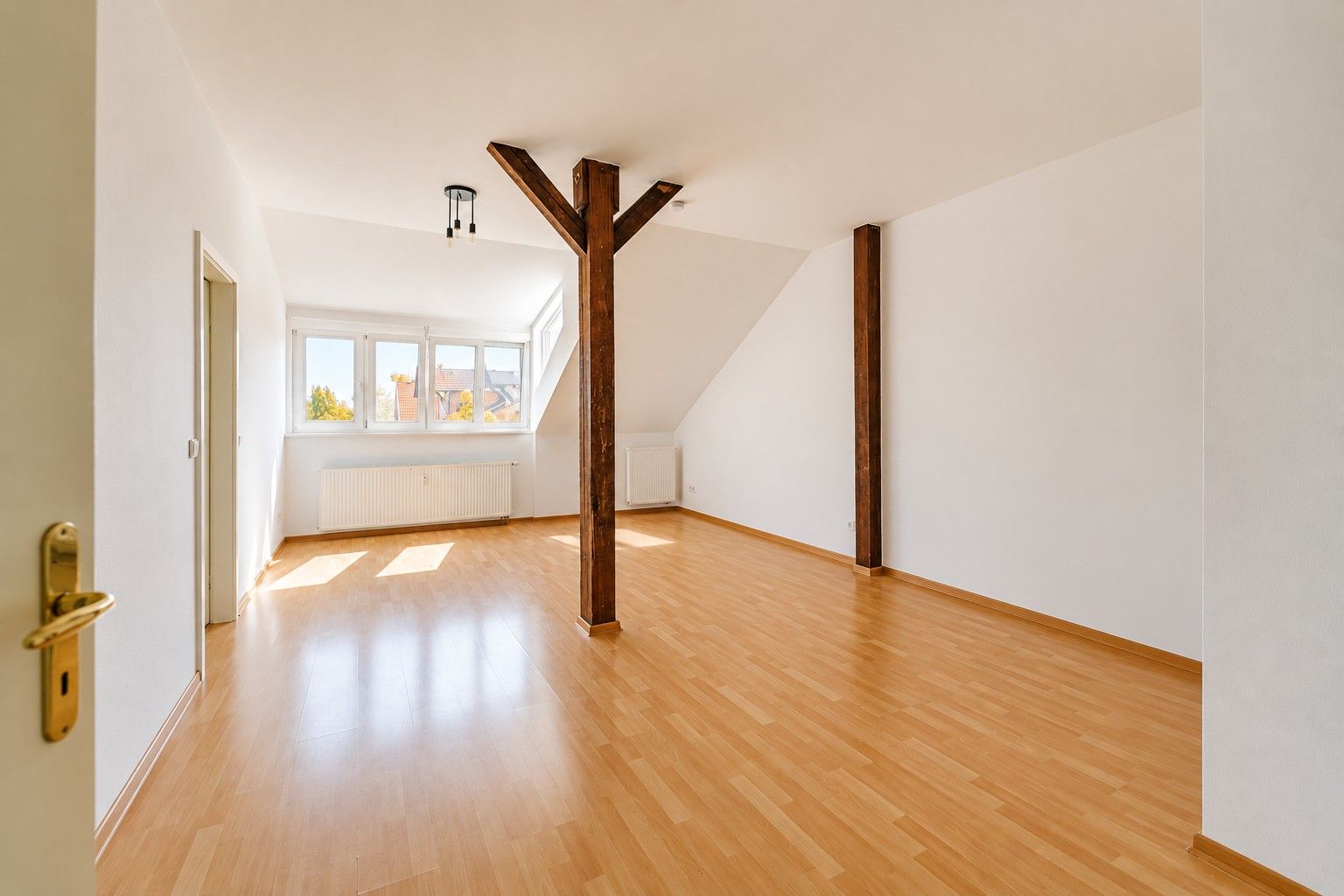 Helles, leeres Zimmer mit Holzfußboden, weißen Wänden und sichtbaren Holzbalken.