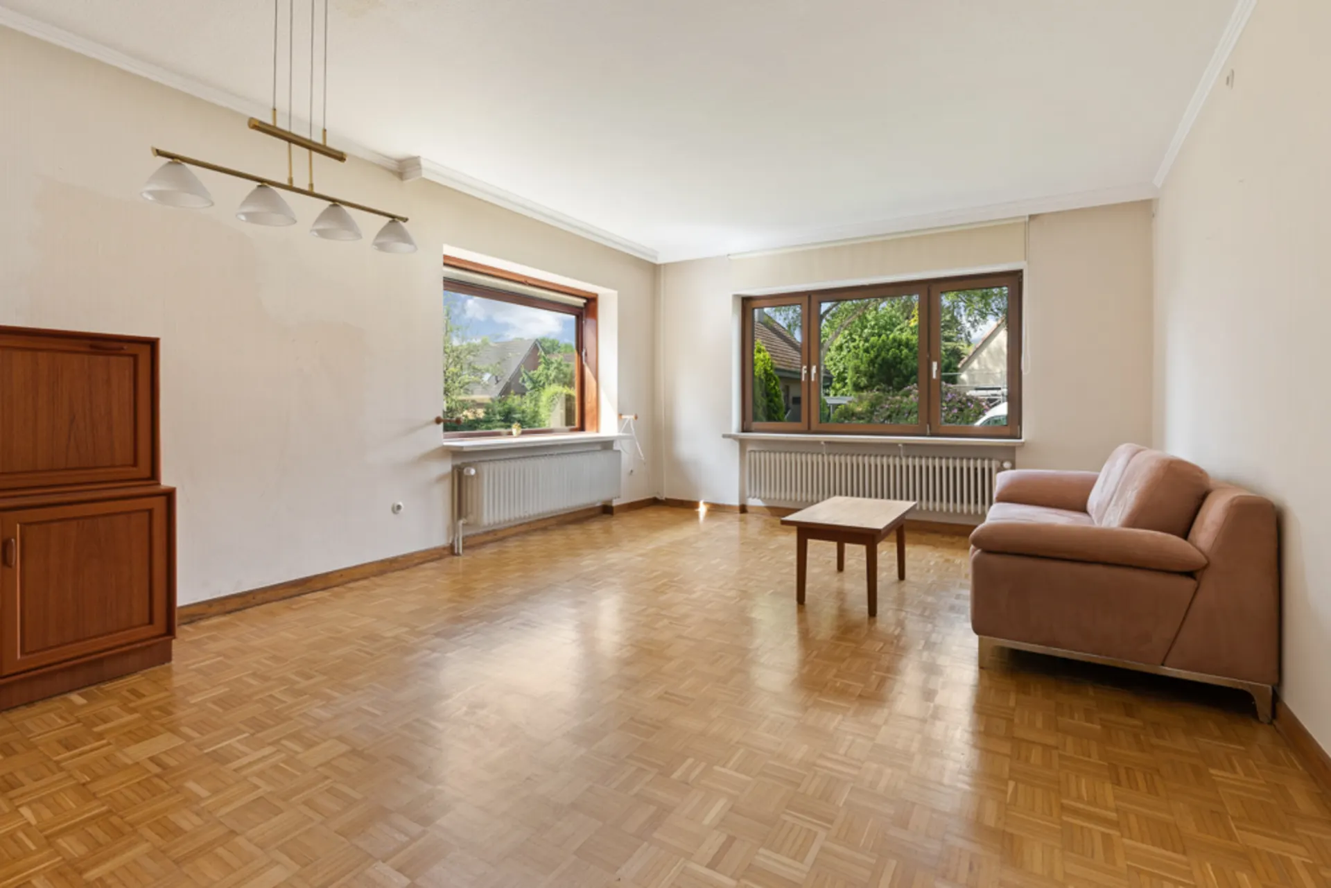 Helles Wohnzimmer mit Parkettboden, braunem Sofa, Couchtisch, Sideboard und großen Fenstern.