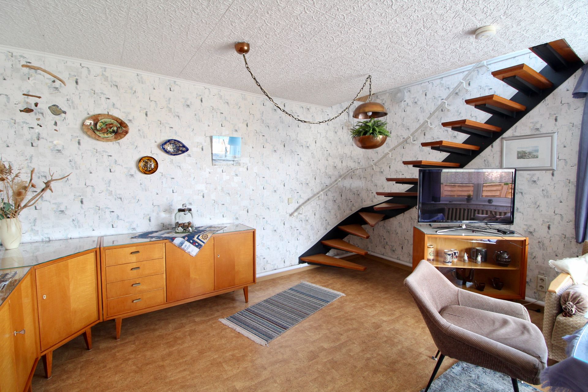 Wohnzimmer mit Holztreppe, Sideboard, Fernseher und Sessel, dekoriert mit Bildern und Pflanzen.
