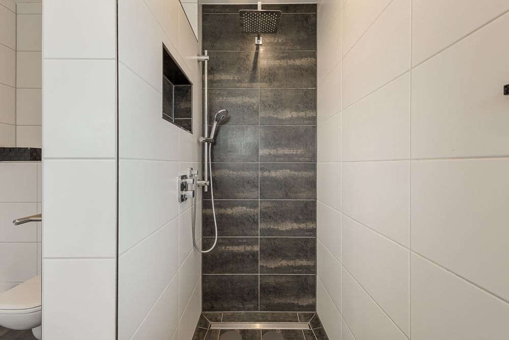 Moderne Dusche mit dunklen Fliesen und Regenduschkopf, daneben WC teilweise sichtbar.