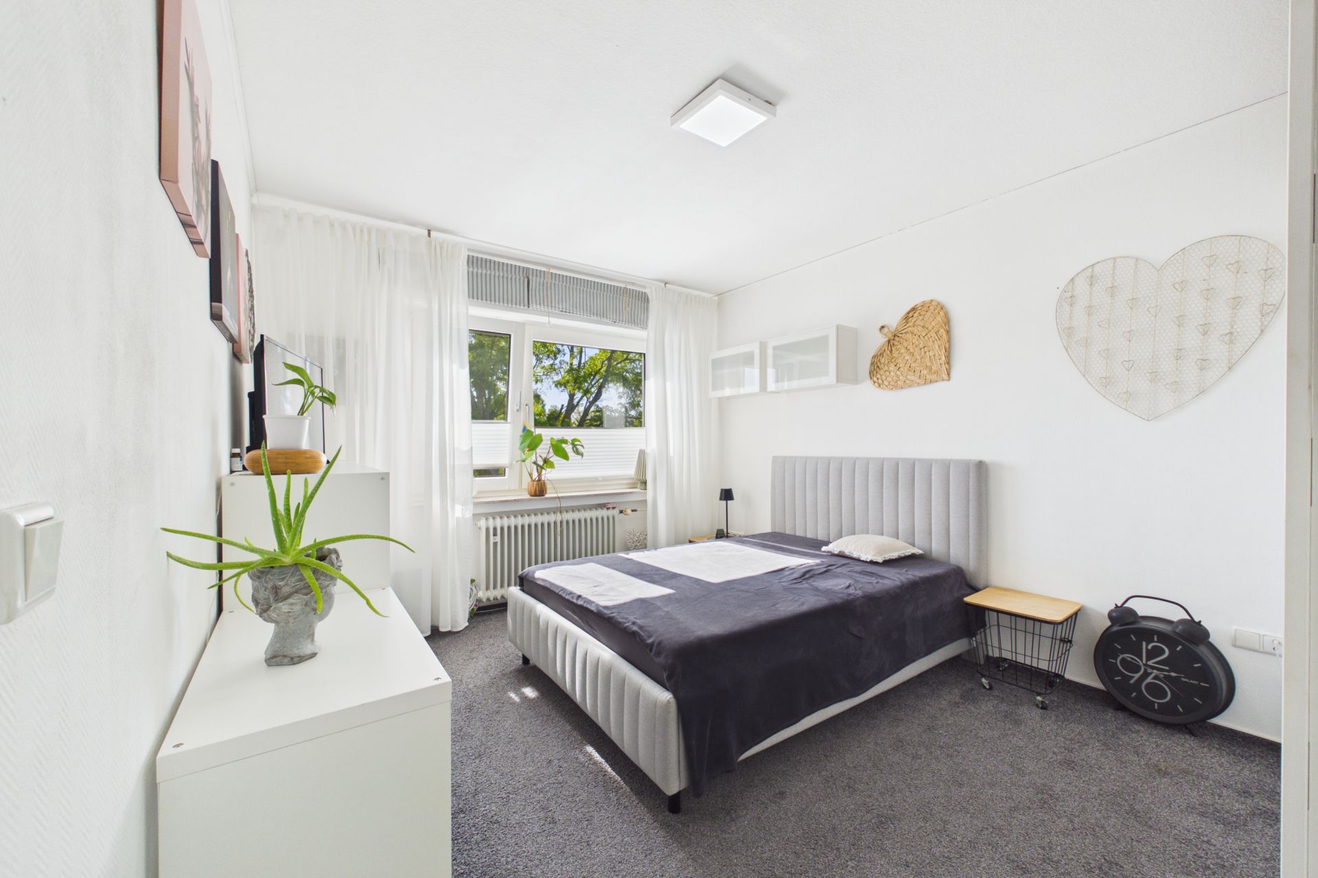 Helles Schlafzimmer mit grauem Teppich, Doppelbett und großen Fenstern mit weißen Vorhängen.