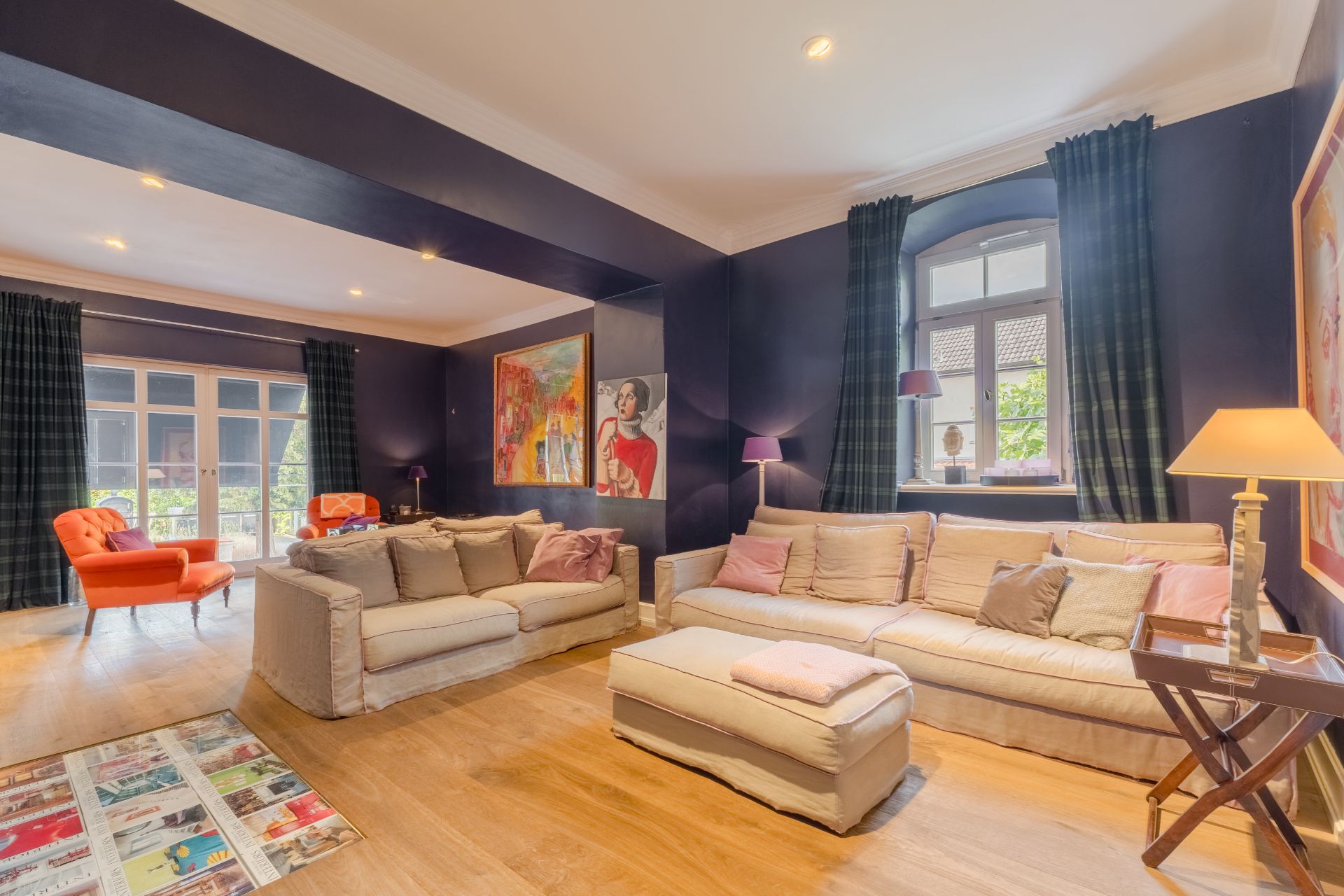 Helles Wohnzimmer mit zwei Sofas, bunten Bildern an der Wand und großen Fenstern mit dunklen Vorhängen.