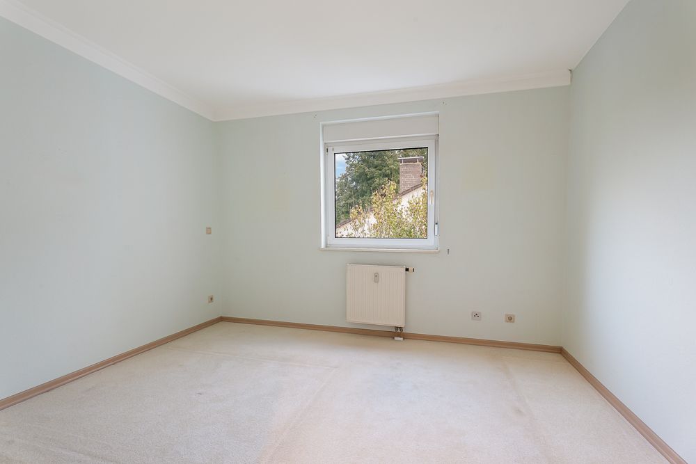 Leeres Zimmer mit cremefarbenem Teppich, hellem Wandanstrich und Fenster mit Blick nach draußen.
