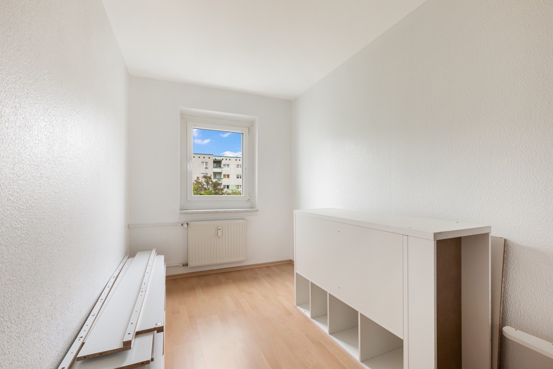 Helles, schmales Zimmer mit Fenster, Heizkörper und einem weißen Regal auf hellem Holzboden.