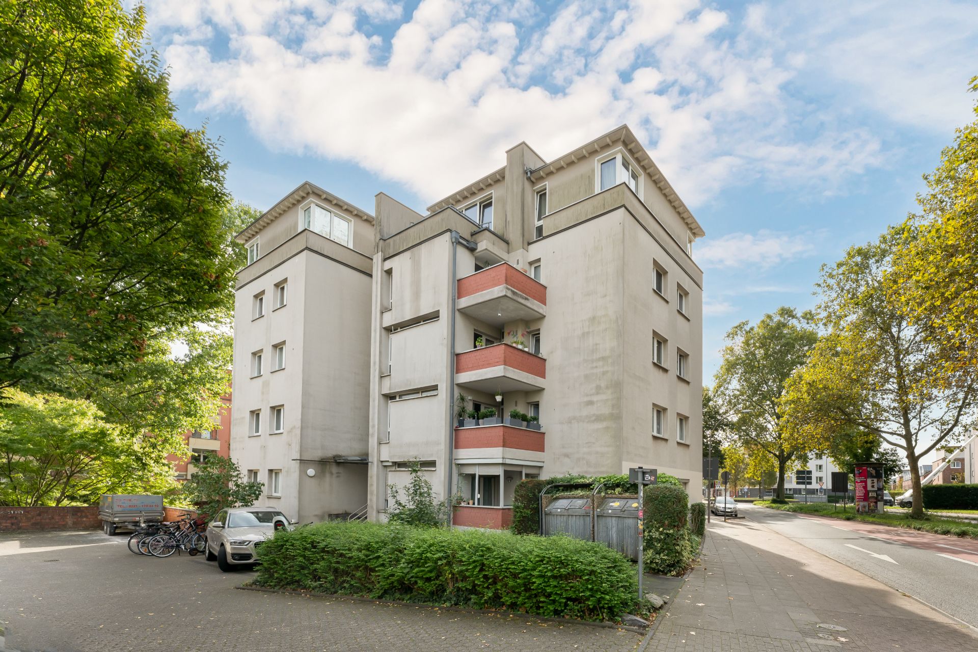 Mehrstöckiges Wohnhaus mit Balkonen, umgeben von Bäumen und geparkten Autos.