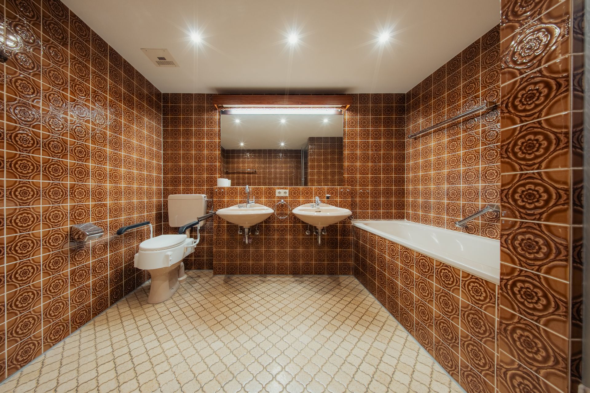 Badezimmer mit braunen Fliesen, zwei Waschbecken, Toilette, Badewanne und großem Spiegel.