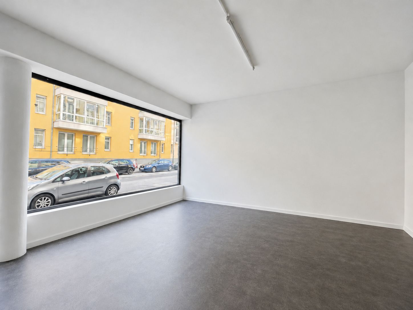 Leerer Raum mit großem Fenster, Blick auf eine Straße mit geparkten Autos und gelbem Wohngebäude.