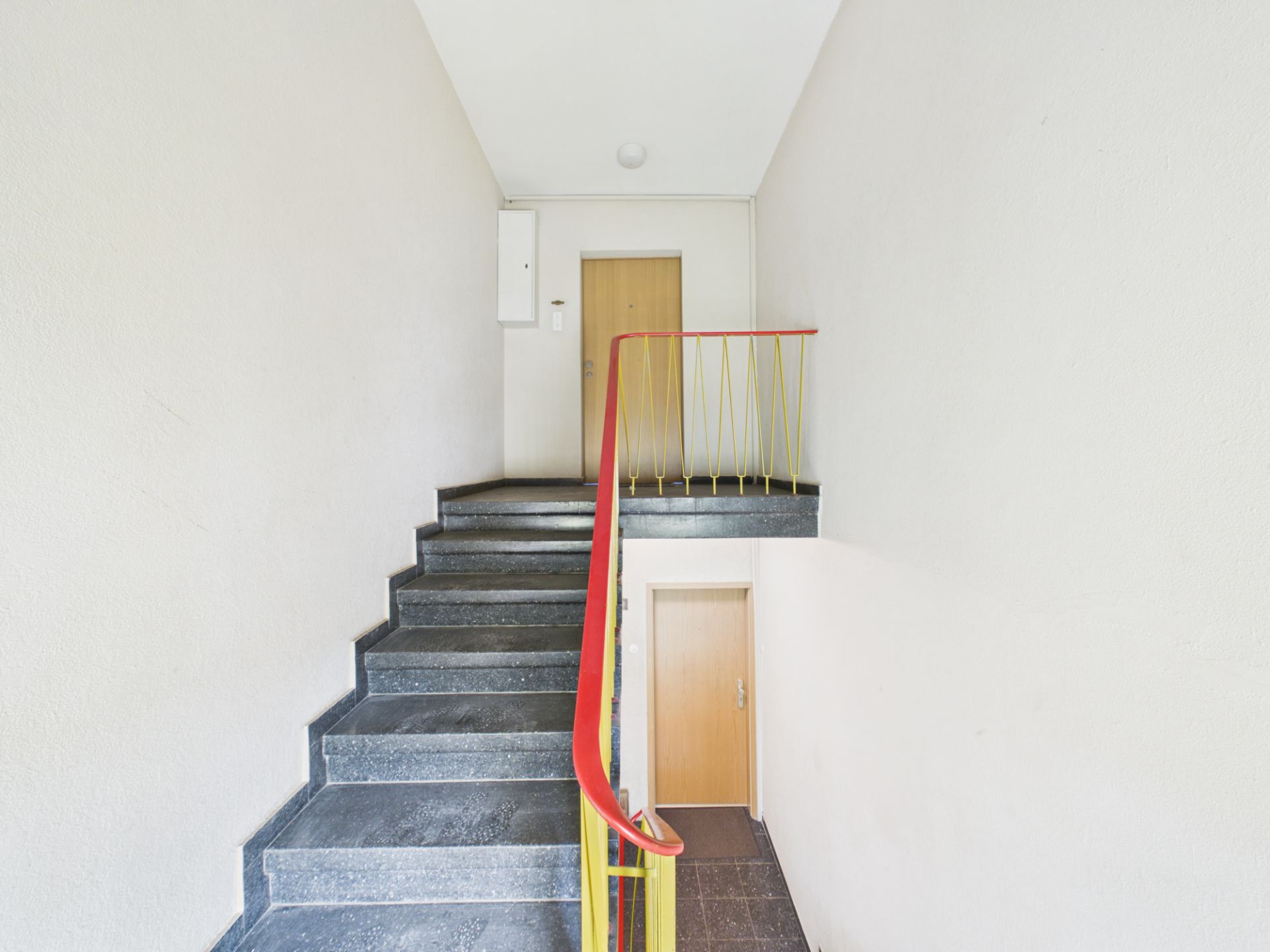 Treppenhaus mit grauer Treppe, rotem Geländer und zwei Holztüren auf verschiedenen Ebenen.