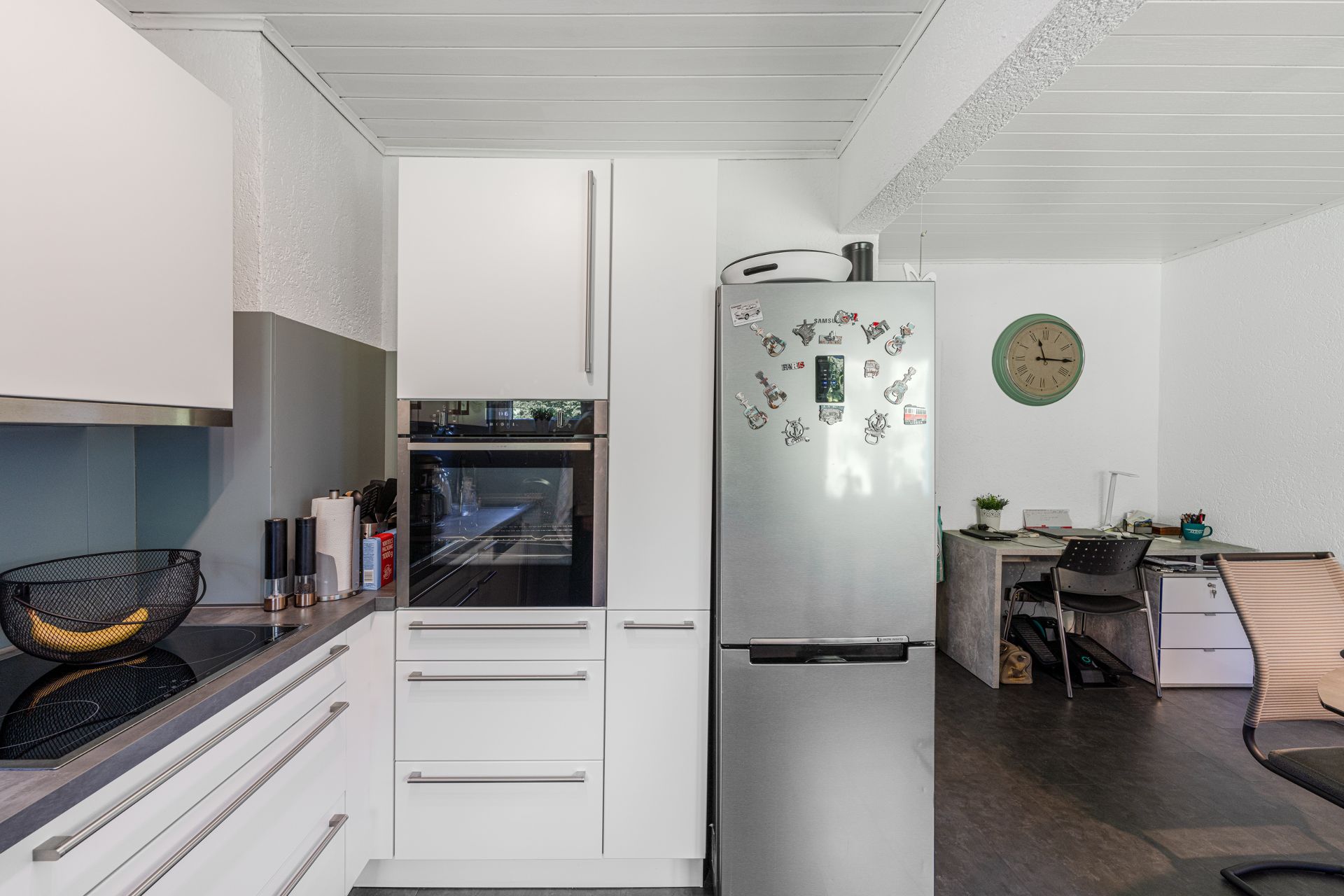 Moderne Küche mit weißem Kühlschrank und Schreibtischecke mit Stuhl und grüner Wanduhr.