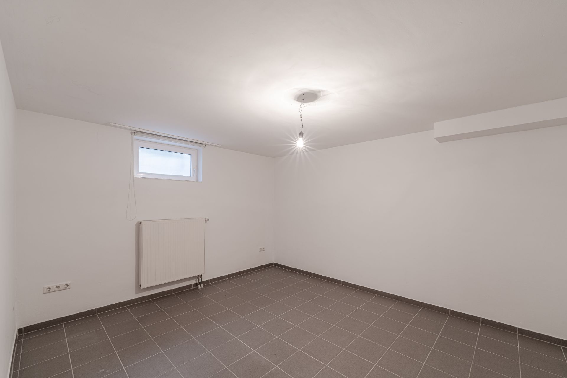 Leerer Raum mit grauen Fliesen, weiß gestrichenen Wänden, kleinem Fenster und Heizkörper.
