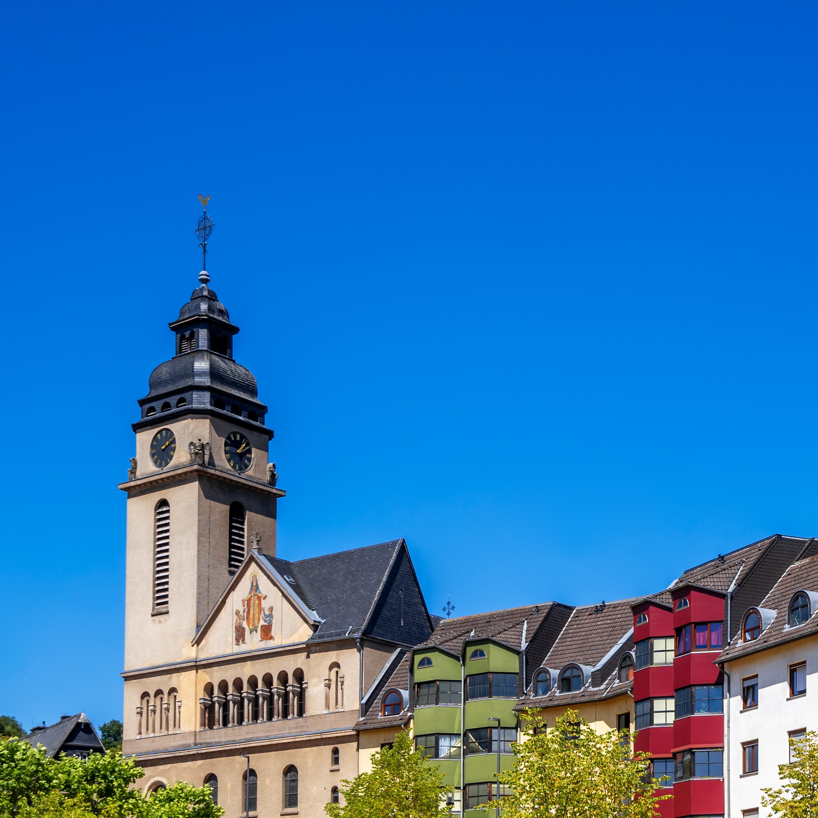 Kirchturm mit Uhr und bunte Wohngebäude im Vordergrund, blauer Himmel.