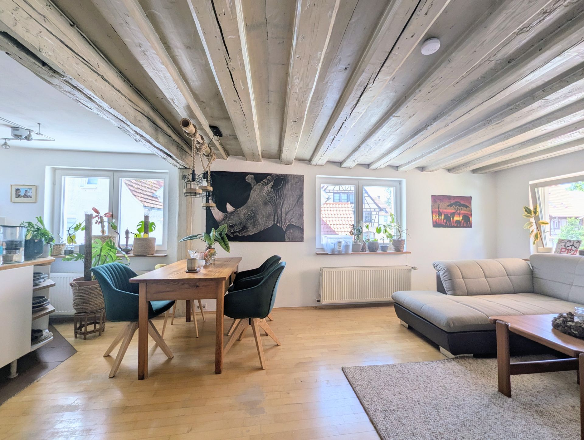 Helles Wohnzimmer mit Holzbalkendecke, Esstisch, Sofa und vielen Pflanzen vor mehreren Fenstern.