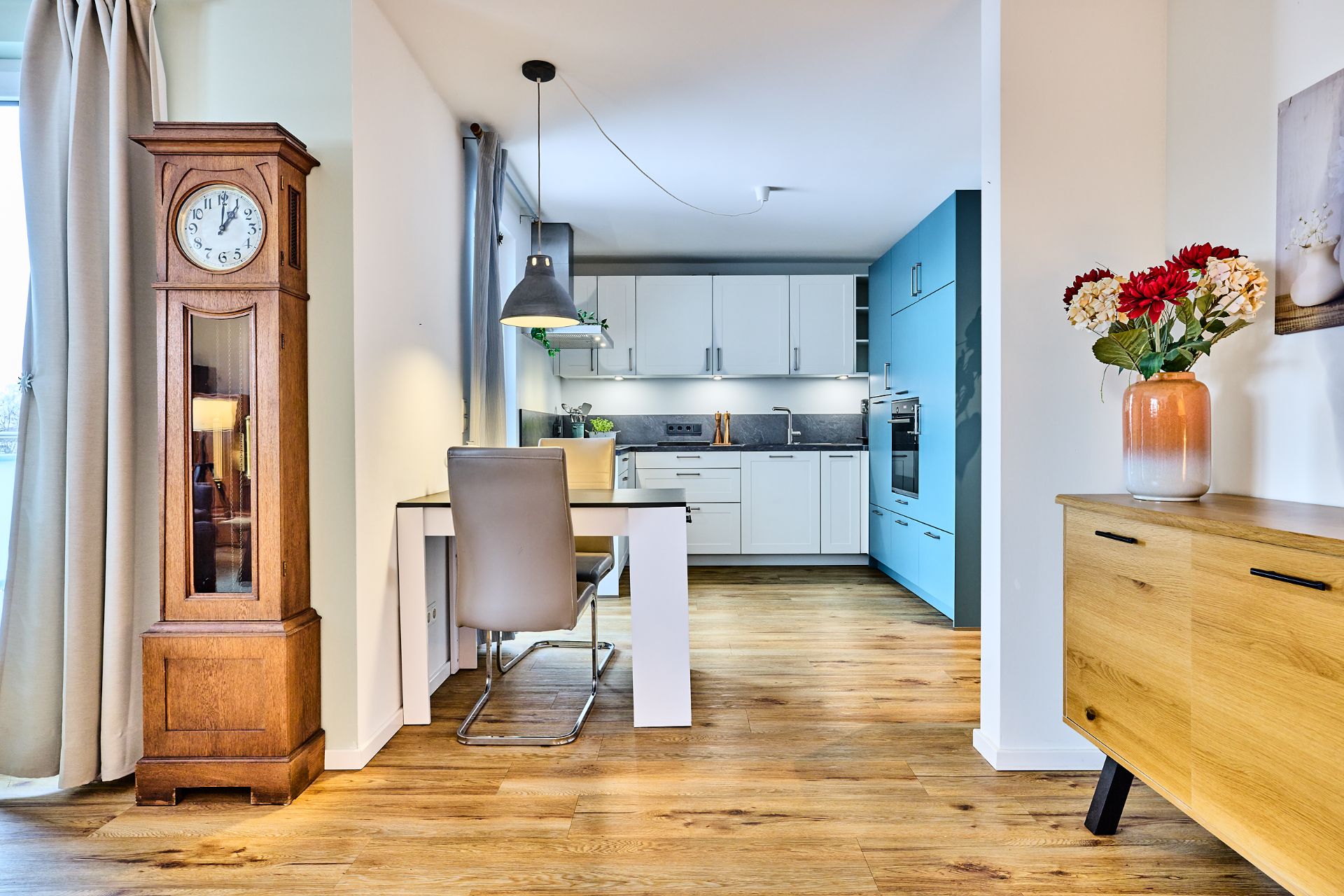Moderne Küche mit weißem Tisch, Stuhl, Standuhr und Kommode mit Blumen auf Holzfußboden.