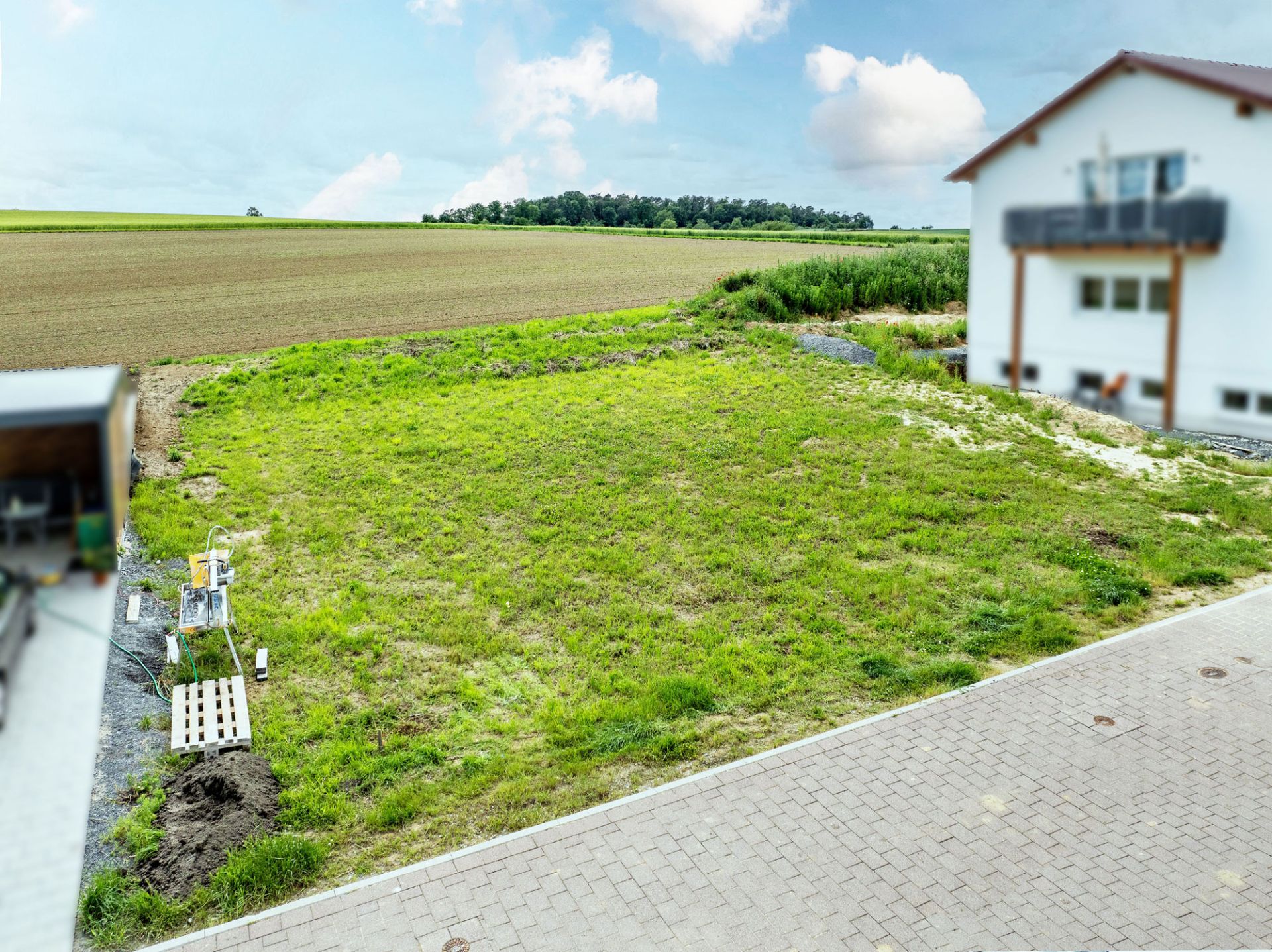 Grünes Grundstück neben gepflasterter Straße, angrenzend an ein weißes Wohnhaus und Felder.